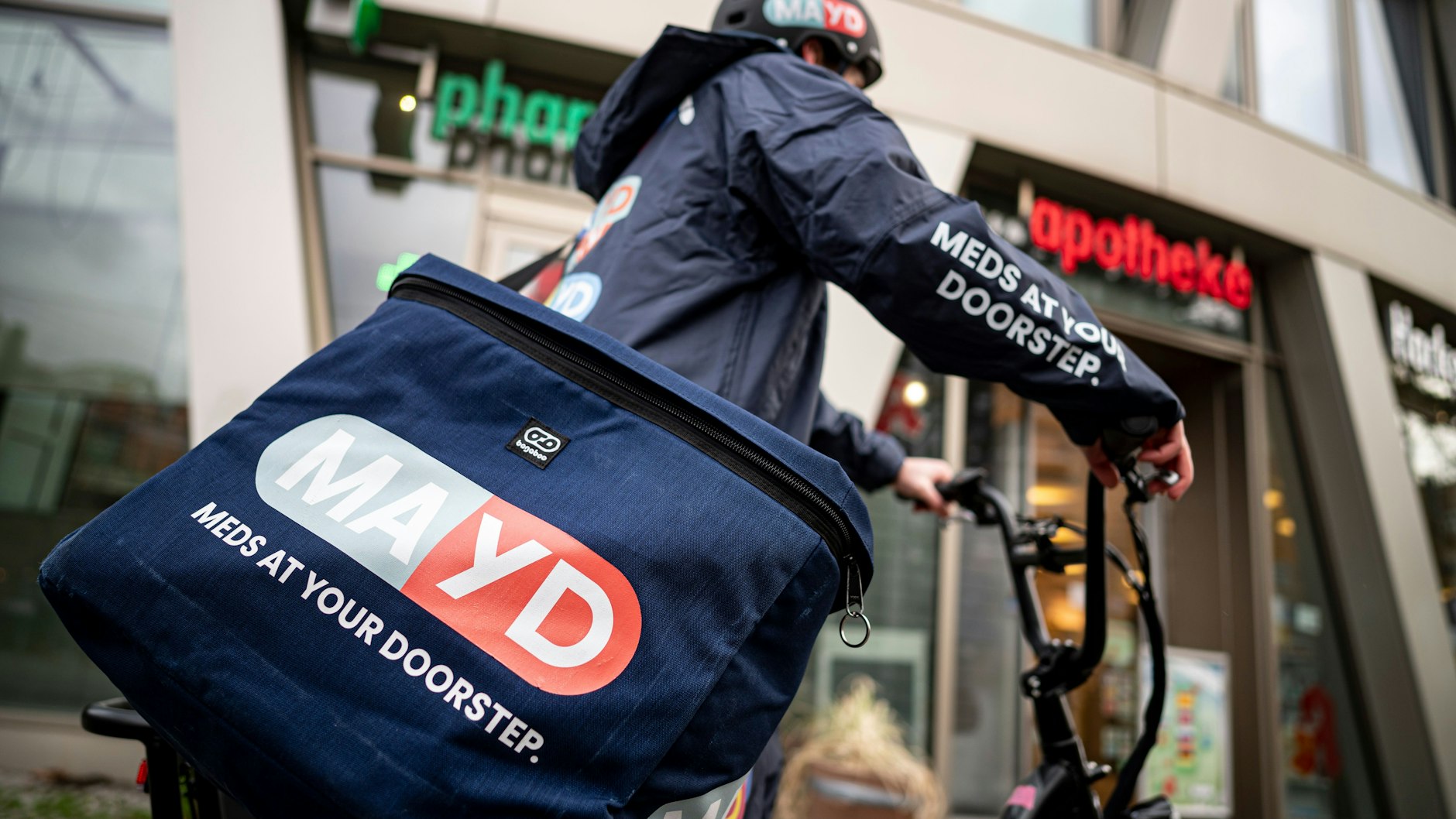 Ein Fahrer des Medikamenten-Lieferservice Mayd steht vor einer Apotheke. Per Mayd-App können Patienten rezeptfreie Medikamente bei ihrer Apotheke vor Ort bestellen. Ein Fahrer oder eine Fahrerin des Start-ups holt die Bestellung dort ab und liefert sie binnen 30 Minuten nach Hause - auch nach Ladenschluss und sonntags.