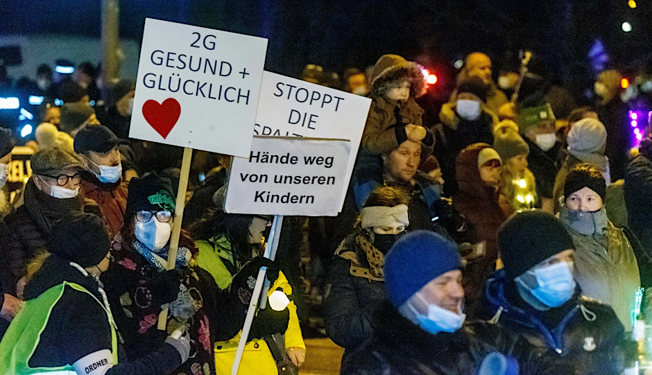 Demonstranten in Lübeck tragen Protestschilder mit Aufschriften wie „2G – Gesund und glücklich“, „Stoppt die Spaltung“ und „Hände weg von unseren Kindern“.