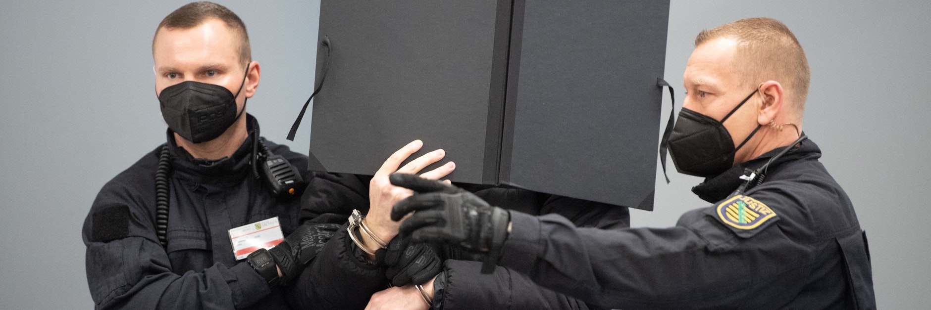 Ein Angeklagter wird mit Handschellen gefesselt von zwei Justizwachtmeistern in den Verhandlungssaal geführt.