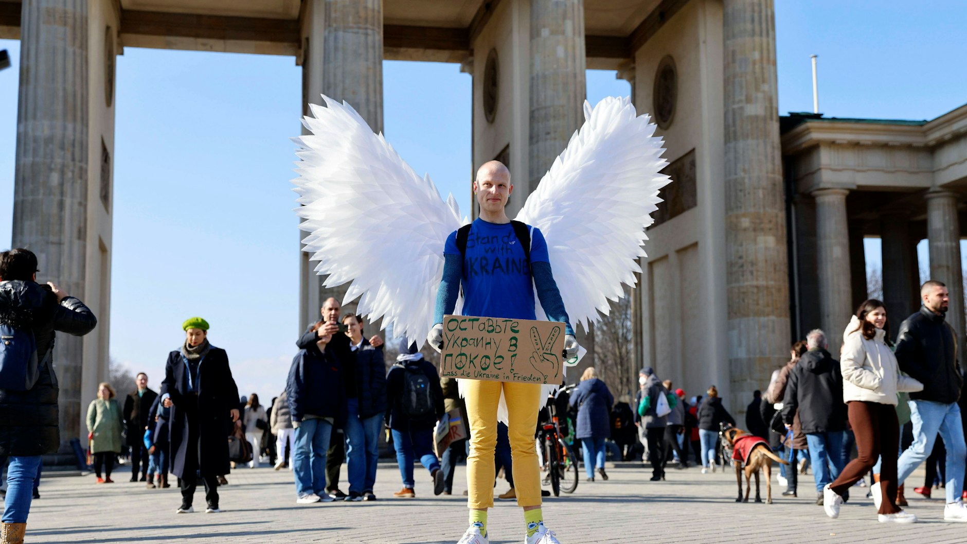 Ein Mann, in die ukrainischen Farben gekleidet, hat sich die Flügel eines Friedensengels umgehängt. Auf seinem Pappschild steht „Lass die Ukraine in Frieden“.