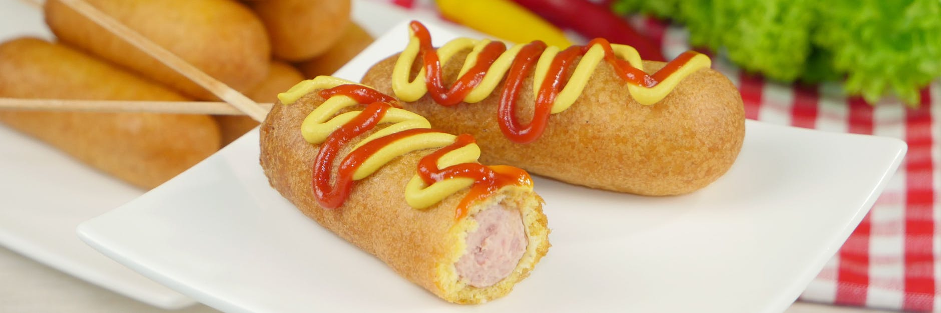 Klassische Corn Dogs bestehen aus aufgespießten Hot-Dog-Würstchen in einer Maisteighülle. Traditionell gibt es Senf und Ketchup dazu.
