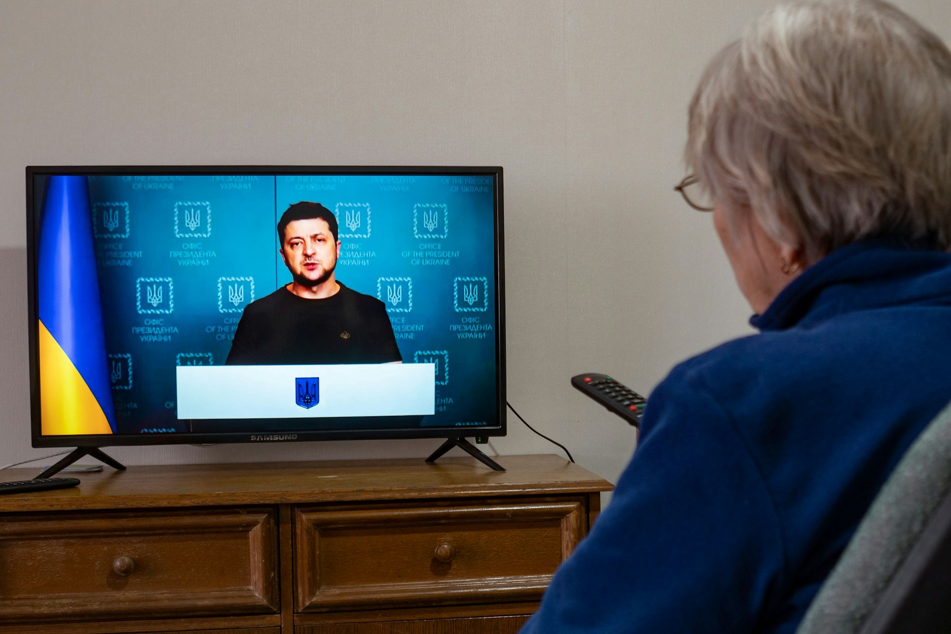 Ein Frau schaut im Fernsehen eine Rede des ukrainischen Präsidenten <a href="https://www.google.com/search?rlz=1C1GCEA_enDE921DE922&amp;q=Wolodymyr+Selenskyj&amp;stick=H4sIAAAAAAAAAONgVuLSz9U3KDfJMqoyfMRoyi3w8sc9YSmdSWtOXmNU4-IKzsgvd80rySypFJLgYoOy-KR4uJC08SxiFQ7Pz8lPqcytLFIITs1JzSvOrswCAP7eeqZcAAAA&amp;sa=X&amp;ved=2ahUKEwjC3oif4r32AhX-SPEDHRPZCDUQzIcDKAB6BAgTEAE">Wolodymyr Selenskyj</a> an.