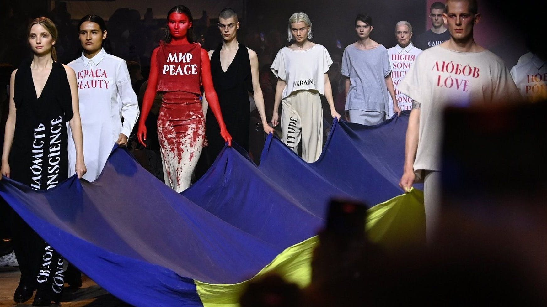 Im Kraftwerk Berlin präsentieren Models Kreationen des ukrainischen Designers Jean Gritsfeldt und tragen dazu eine ukrainische Fahne. Weil er nicht nach Berlin kommen kann und auch seine Kollektion nicht geliefert werden konnte, haben Freiwillige seine Designs nachgeschneidert, damit sie gezeigt werden können.