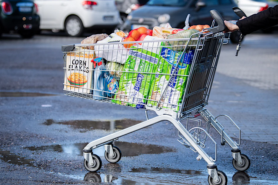  Ein Kunde schiebt einen bis zum Rand gefüllten Einkaufswagen über den Parkplatz eines Supermarktes.