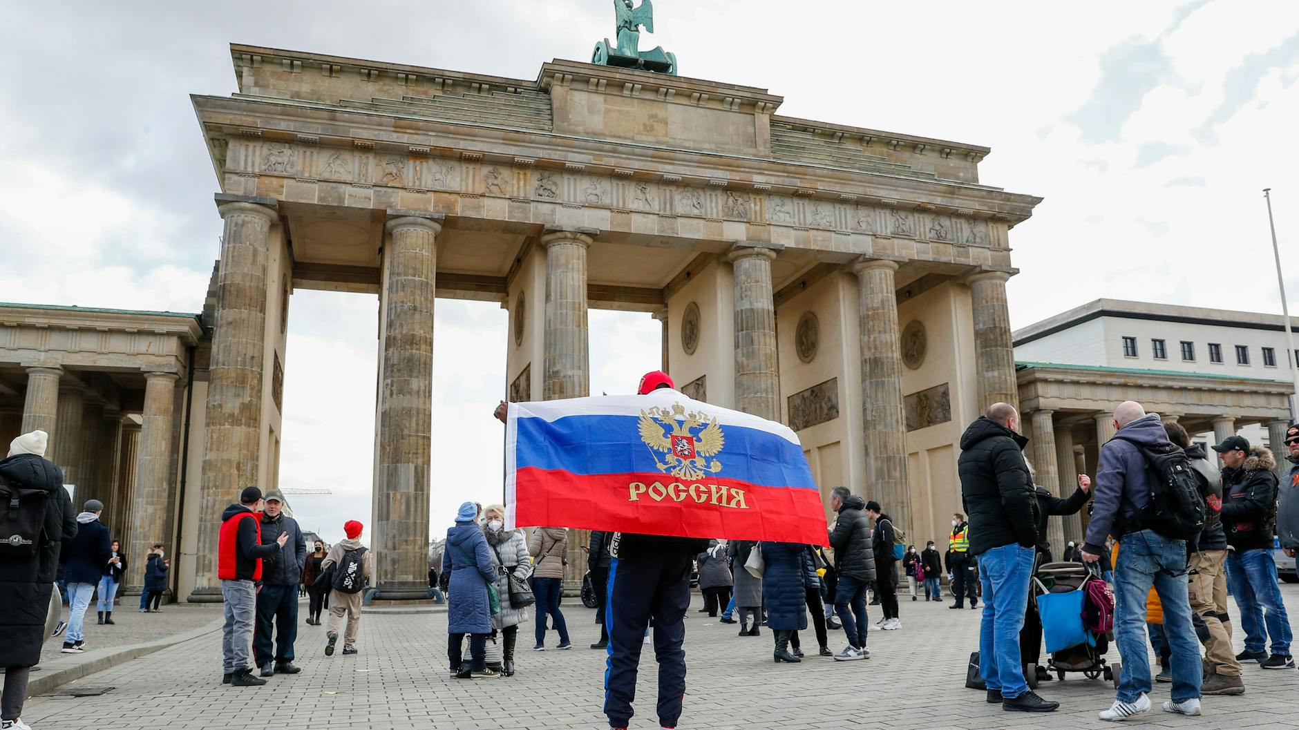 Ein Mann mit einer Flagge Russlands mit Wappen steht vor dem Brandenburger Tor, wo aus Russland stammende und in Deutschland lebende Menschen demonstrieren.