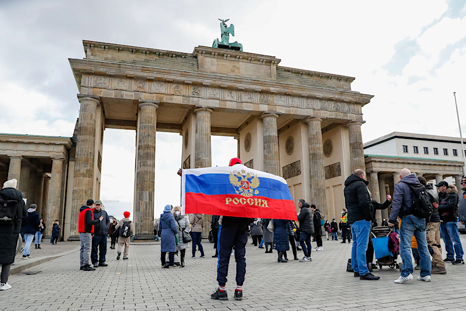 Ein Mann mit einer Flagge Russlands mit Wappen steht vor dem Brandenburger Tor, wo aus Russland stammende und in Deutschland lebende Menschen demonstrieren.