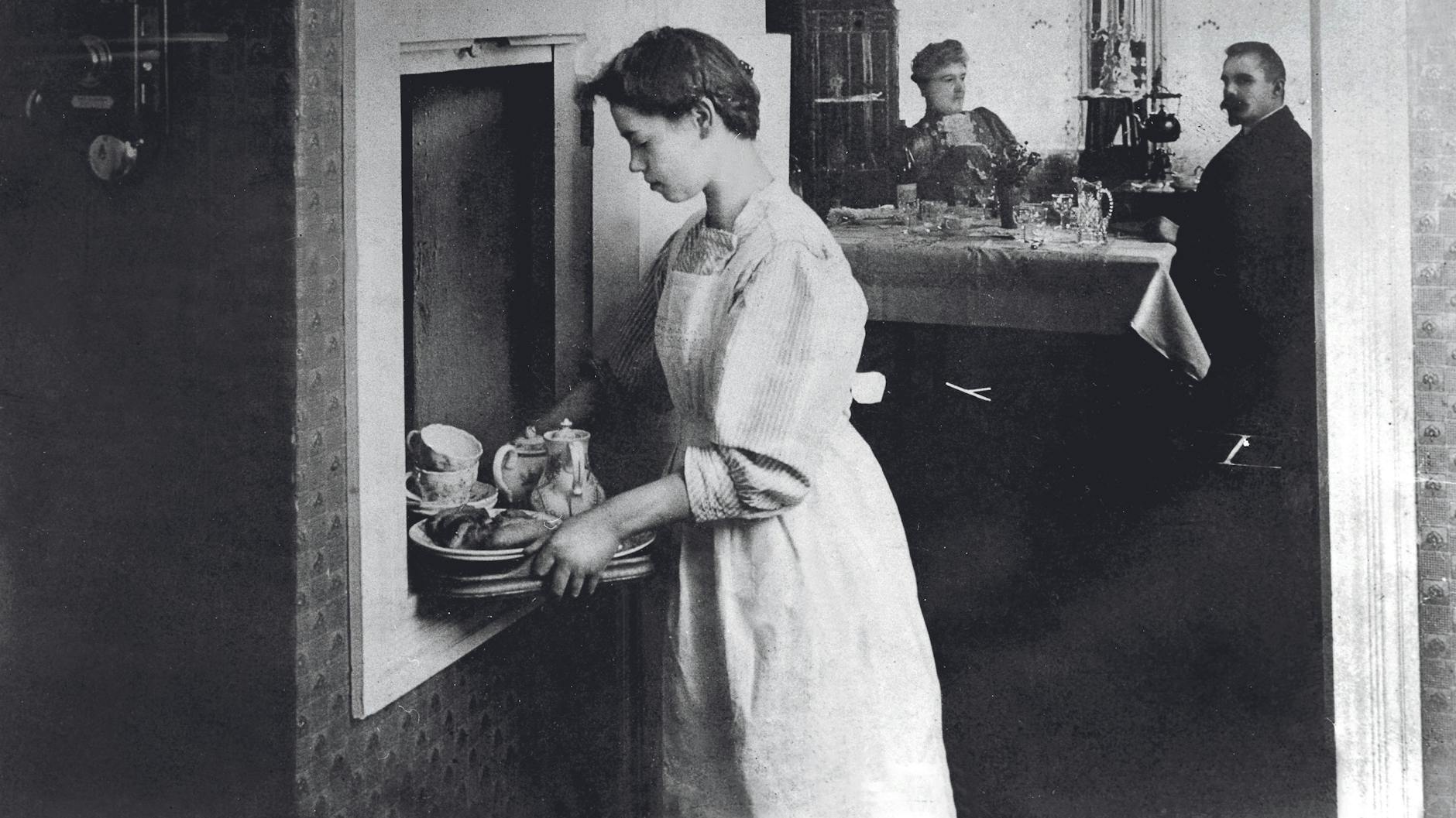 Ein Dienstmädchen serviert seiner Herrschaft eine Mahlzeit aus dem Speisenfahrstuhl.