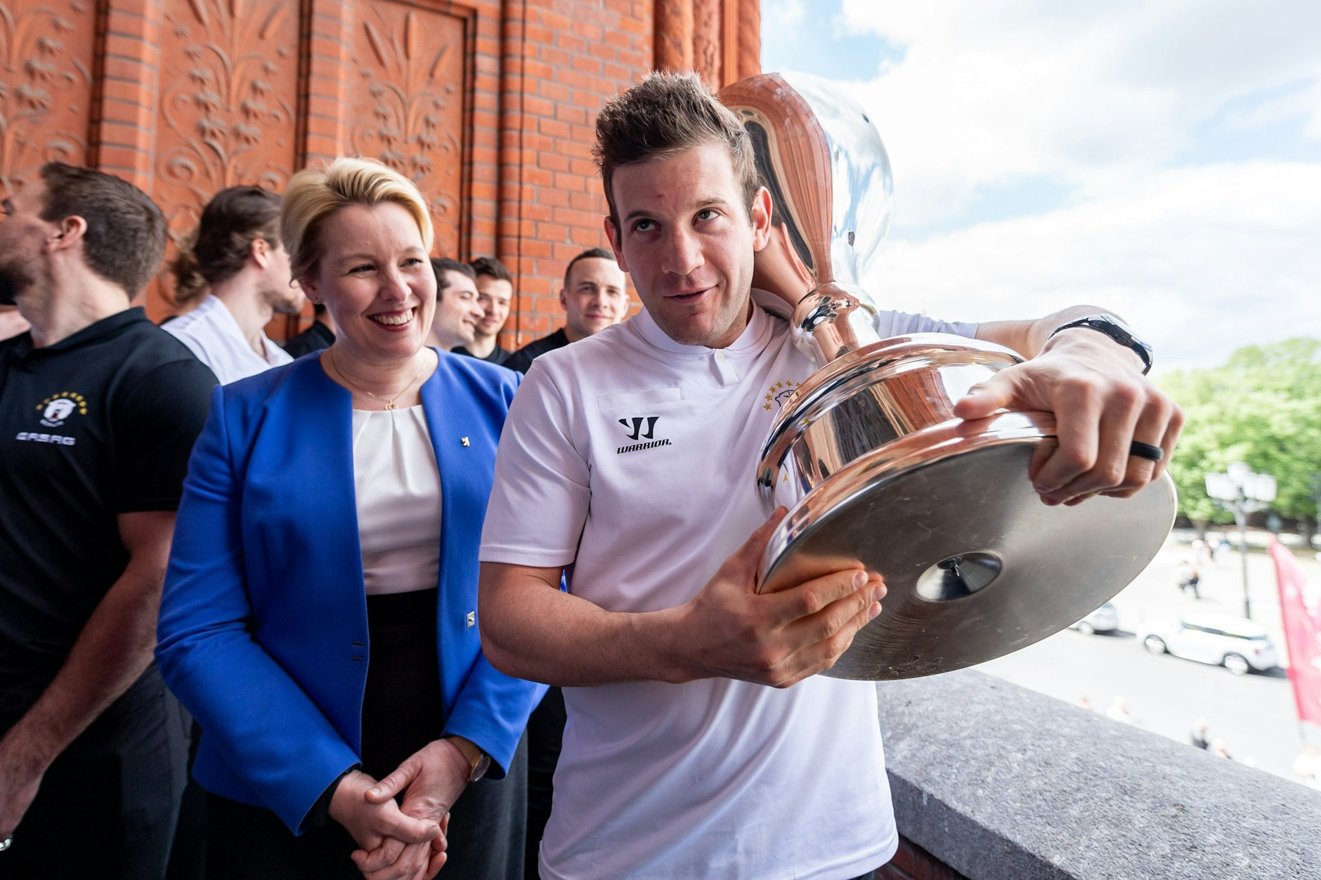 Eisbären-Torjäger Matt White schleppt den Pokal auf dem Rathaus-Balkon, Berlins Regierende Bürgermeisterin Franziska Giffey hat sichtlich Spaß daran.