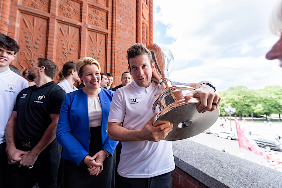 Eisbären-Torjäger Matt White schleppt den Pokal auf dem Rathaus-Balkon, Berlins Regierende Bürgermeisterin Franziska Giffey hat sichtlich Spaß daran.