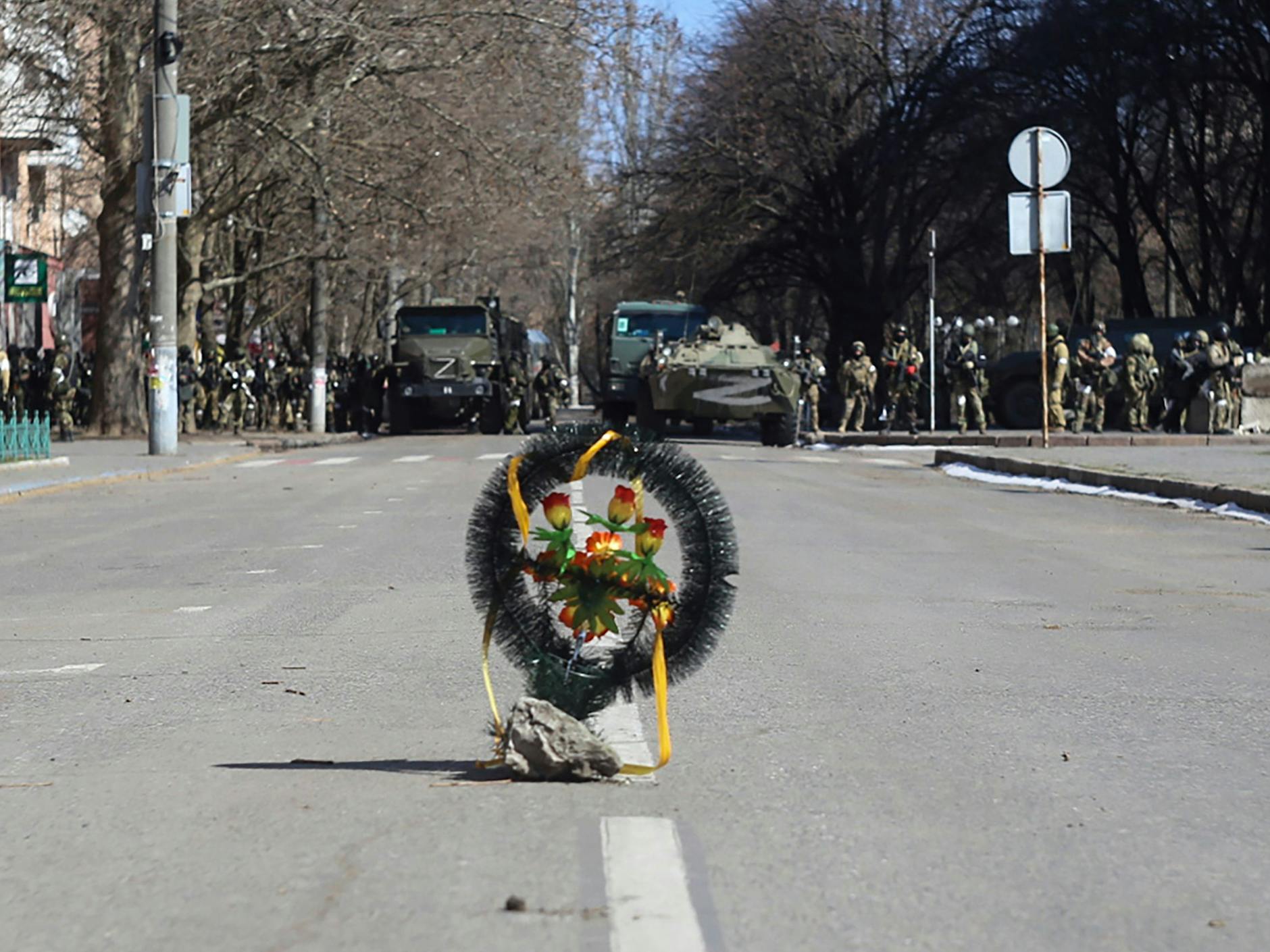 Ein Kranz mit Blumen steht in Cherson auf einer Straße, während Soldaten der russischen Armee neben ihren Lastwagen stehen.