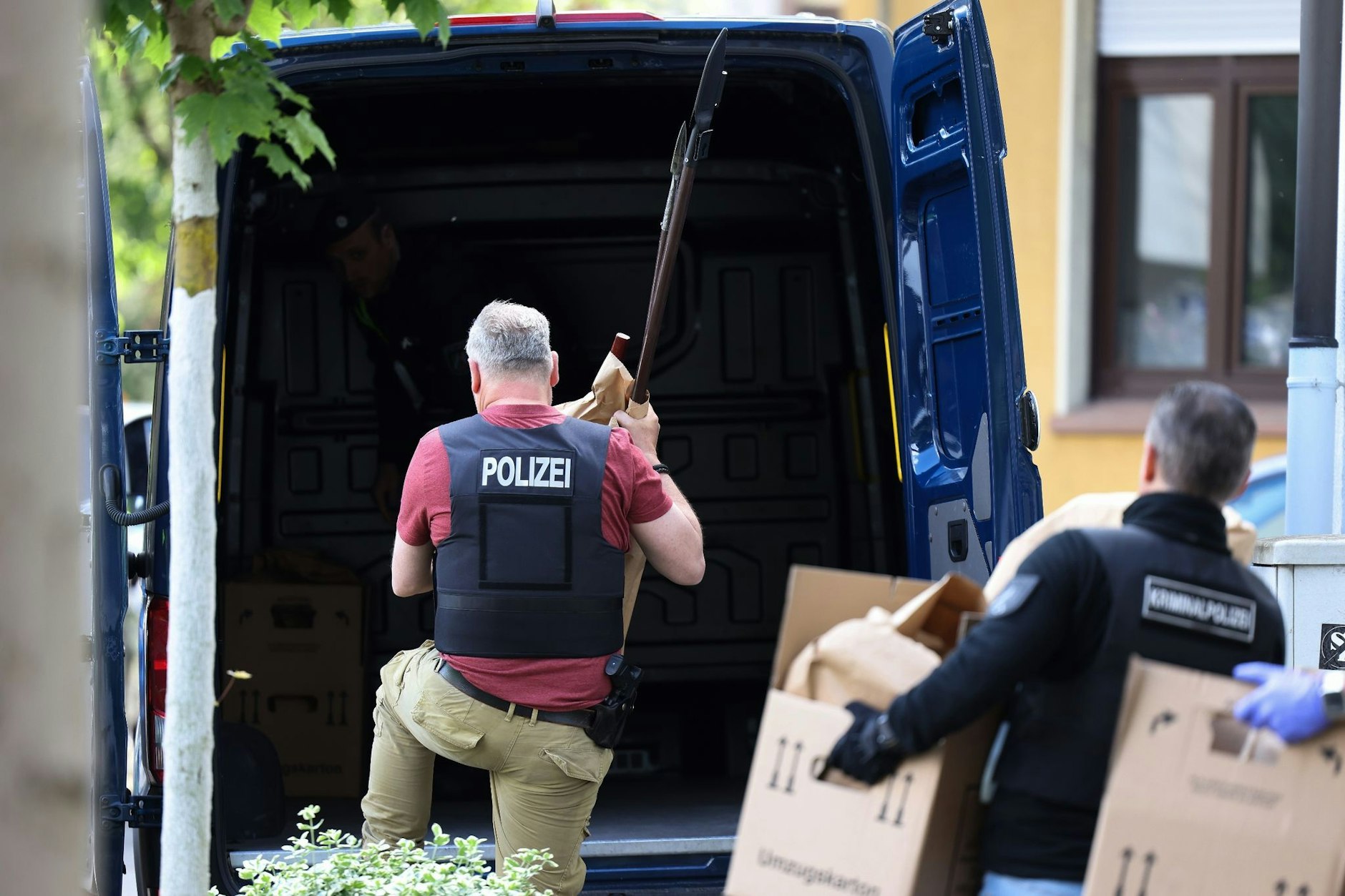 Polizisten tragen Gegenstände, darunter mehrere Stichwaffen und Speere, aus dem Wohnhaus des Verdächtigen.