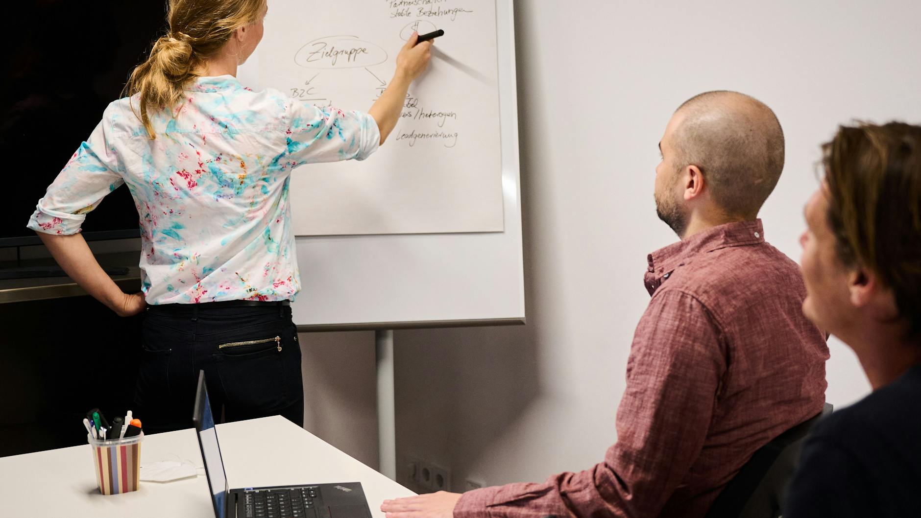 Eine Frau schreibt auf einem Flipchart in einem Meetingraum, während zwei Männer an einem Tisch sitzen (Symbolbild).