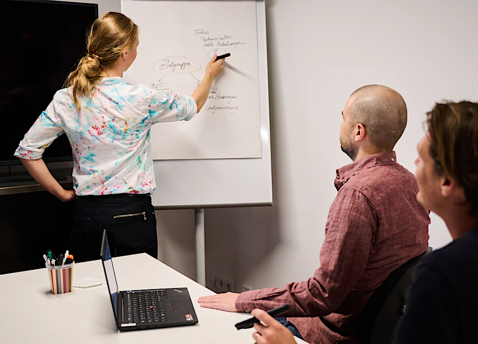 Eine Frau schreibt auf einem Flipchart in einem Meetingraum, während zwei Männer an einem Tisch sitzen (Symbolbild).
