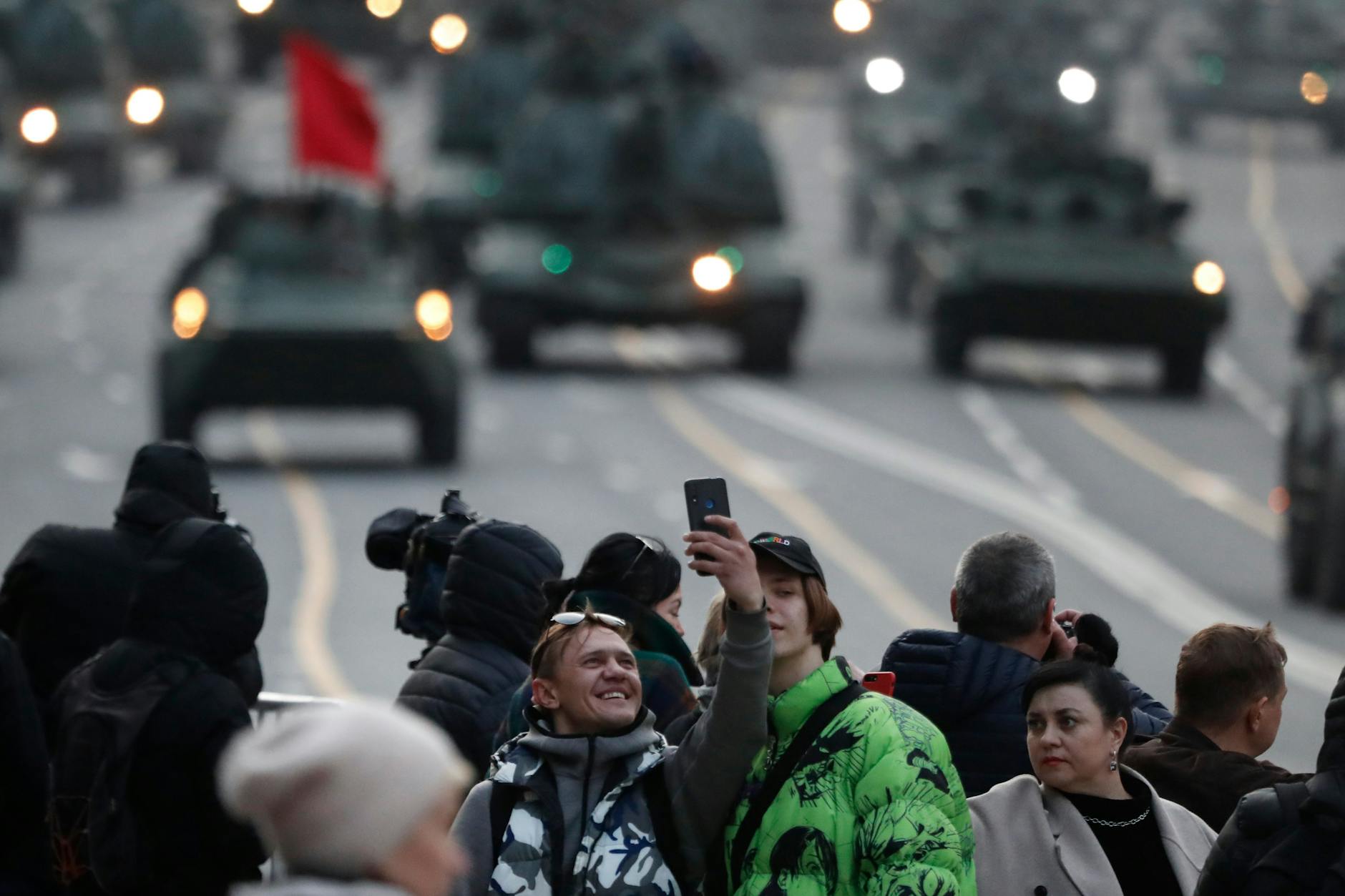 Ein Mann nimmt mit seinem Handy ein Selfie auf während im Hintergrund Fahrzeuge des russischen Militärs anfahren.