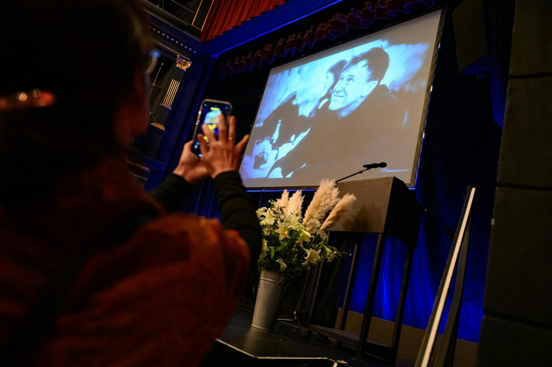 Eine Frau fotografiert mit ihrem Smartphone die Bühne mit Blumenstrauß und einem Foto des Verstorbenen auf einer Leinwand.