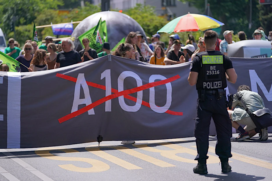 Ein Polizist steht bei einer Demo gegen den Weiterbau der Stadtautobahn A100 vor einem Banner der Demonstranten.  