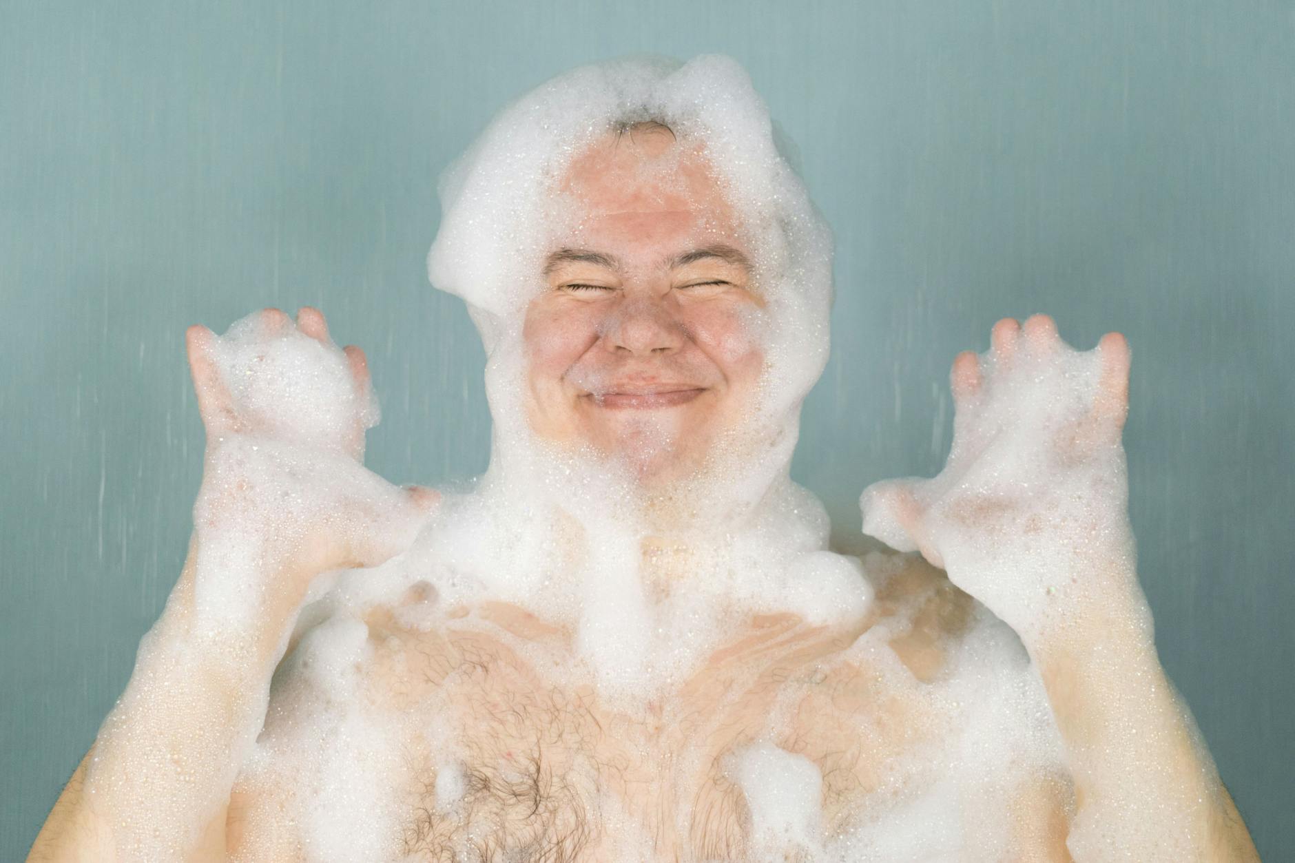 the man squeezes his eyes and smiles in the soapy foam on his head and his hands are lifted, on a green background
