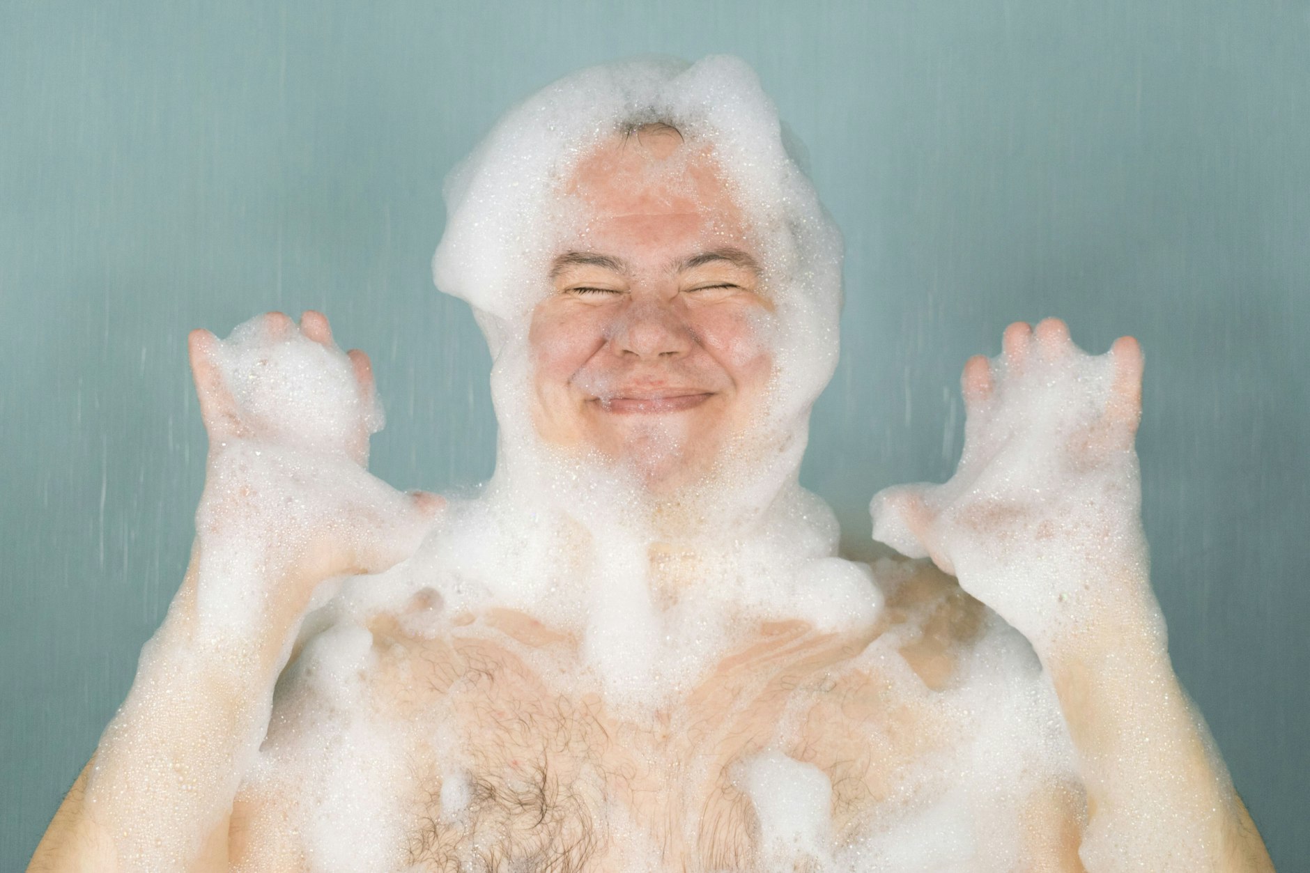 the man squeezes his eyes and smiles in the soapy foam on his head and his hands are lifted, on a green background