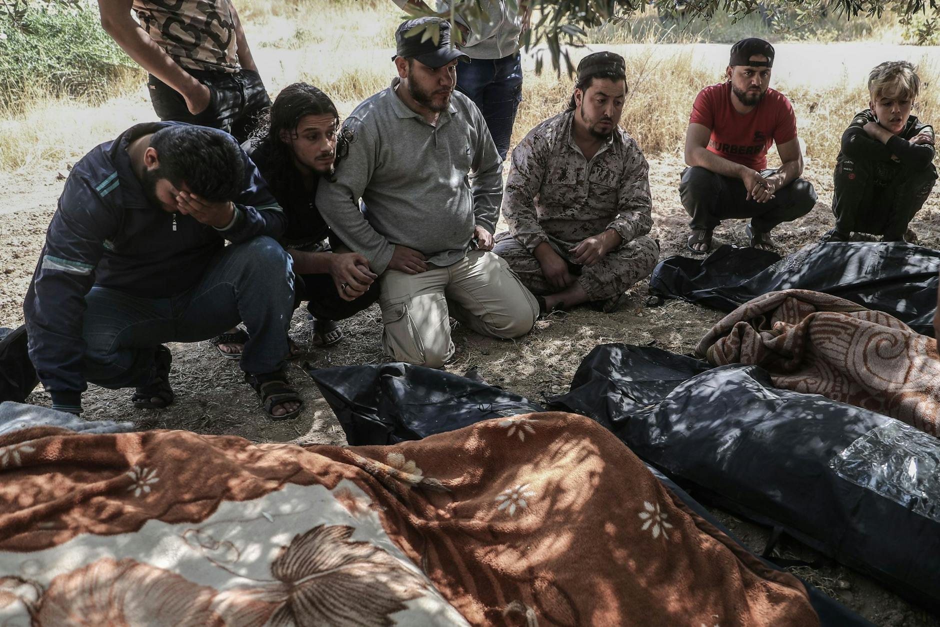 ARCHIV - Trauernde sitzen 2021 neben eingehüllten Leichen unter Bäumen in Idlib und warten darauf, dass Kampfflugzeuge abziehen.