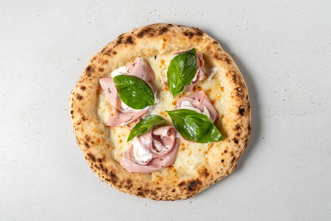 Die sommerliche Pizza ist wunderschön, die Farben wie ein Gemälde: Ein dicker, von Beige ins Dunkelgold spielende Rand, klar neapolitanisch geschwollen mit einigen Blasen.