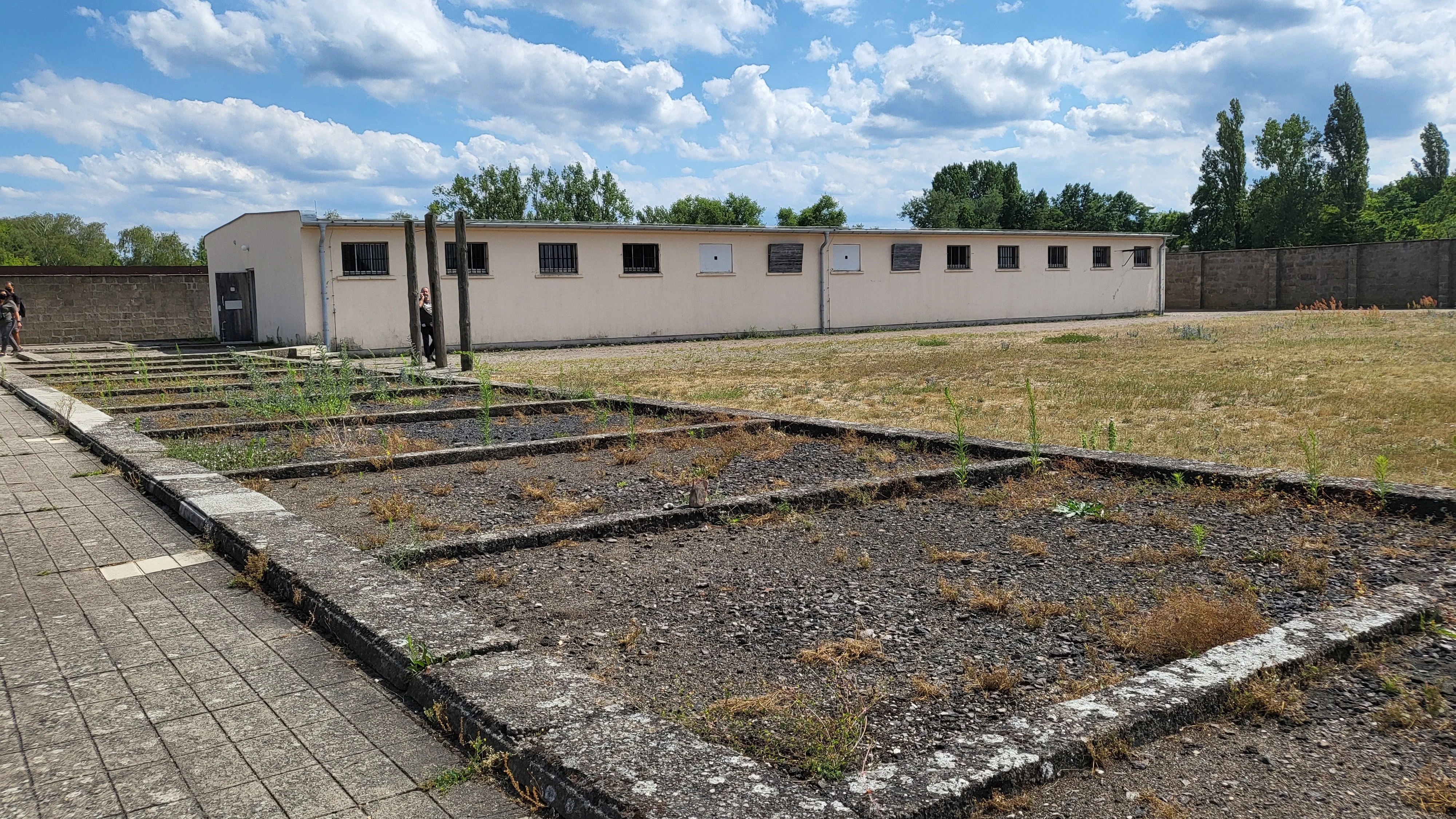 Die Grundmauern von Flügel B des Zellenbaus im KZ Sachsenhausen, am Ort der Zellen 72 und 73, die für Stepan Bandera zusammengelegt wurden. Sie lagen rechts und links der mit einem Stein markierten Grundmauer. Im Hintergrund Flügel A, davor drei Folterpfähle.