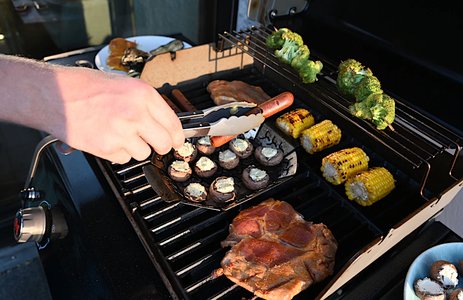 Ein Mann wendet mit einer Zange eine Grillwurst auf einem Gasgrill.
