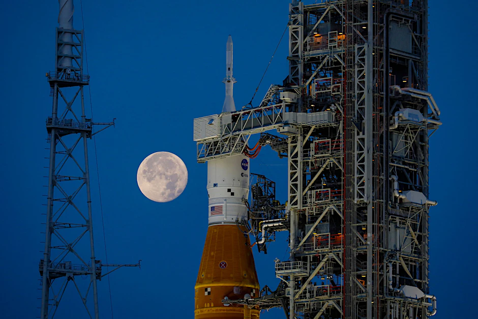 Die Artemis-1 Rakete bei Nacht, am blauen Himmel der Mond. Dort soll es hingehen.