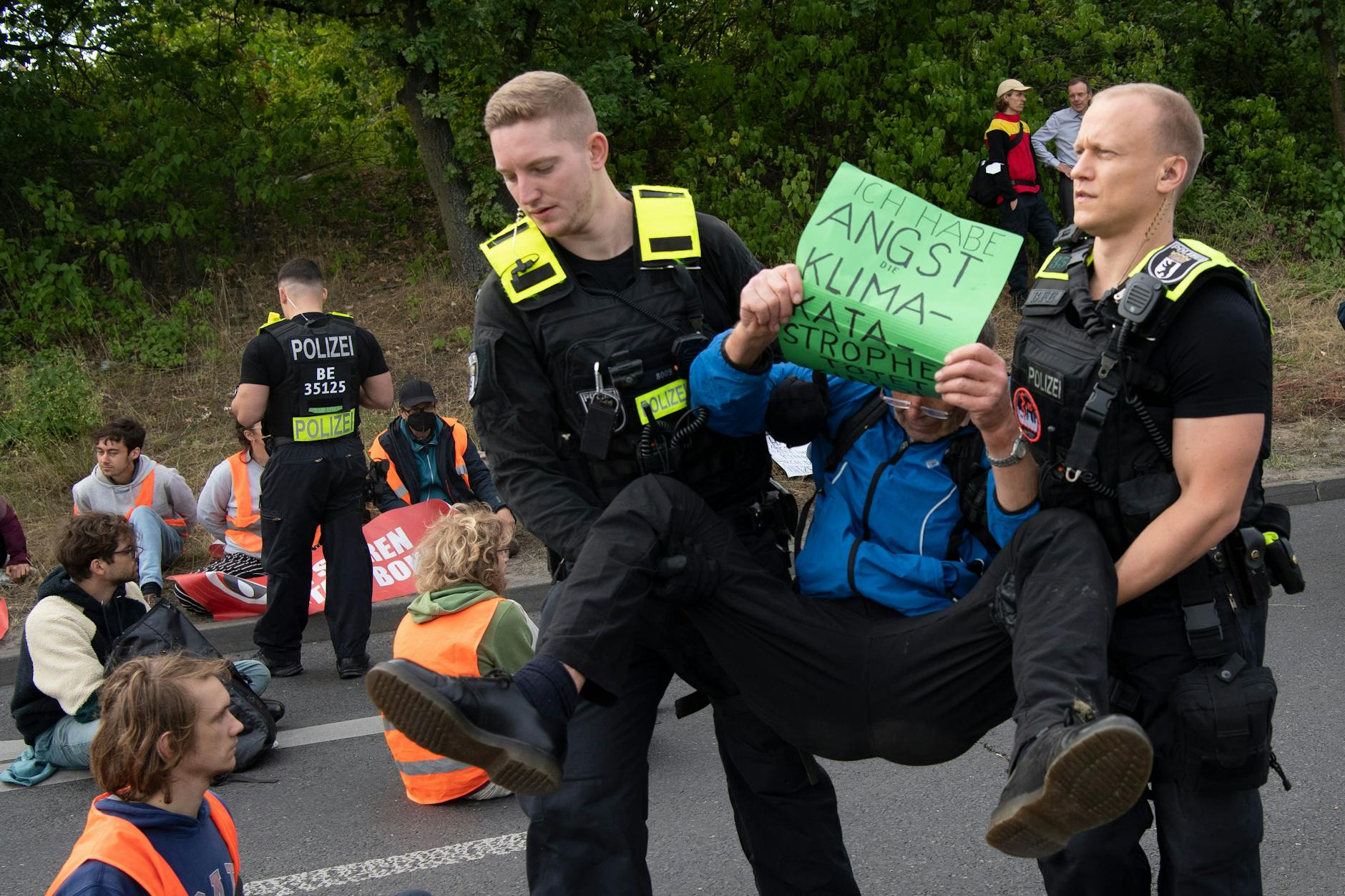 Polizeibeamte tragen einen Demonstranten von der Fahrbahn, der ein Schild mit der Aufschrift „Ich habe Angst Klimakatastrophe tötet Kinder“ vor sich hält.