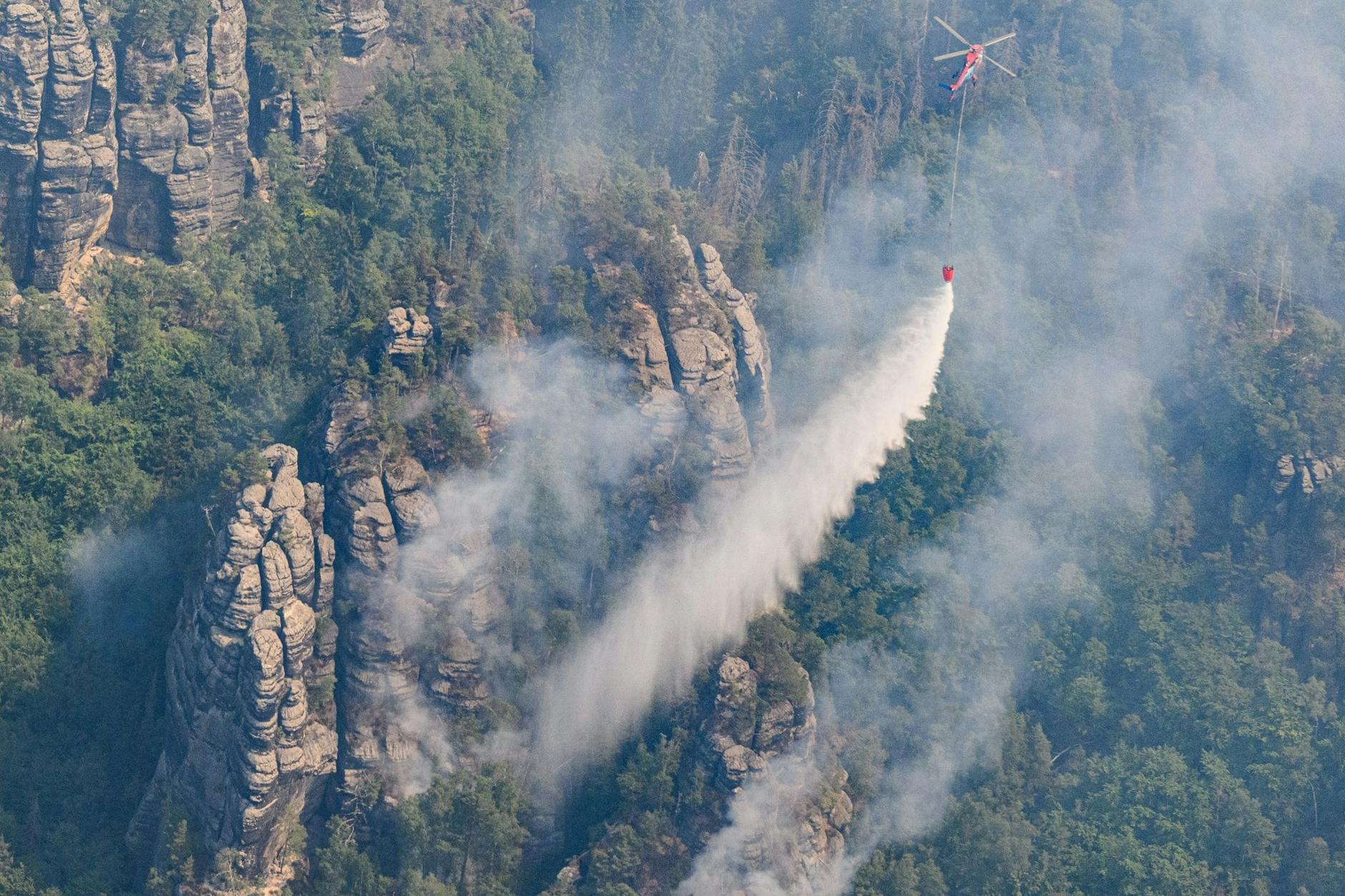Ein Lastenhubschrauber wirft Löschwasser über einem Wald im Nationalpark Sächsische Schweiz ab.