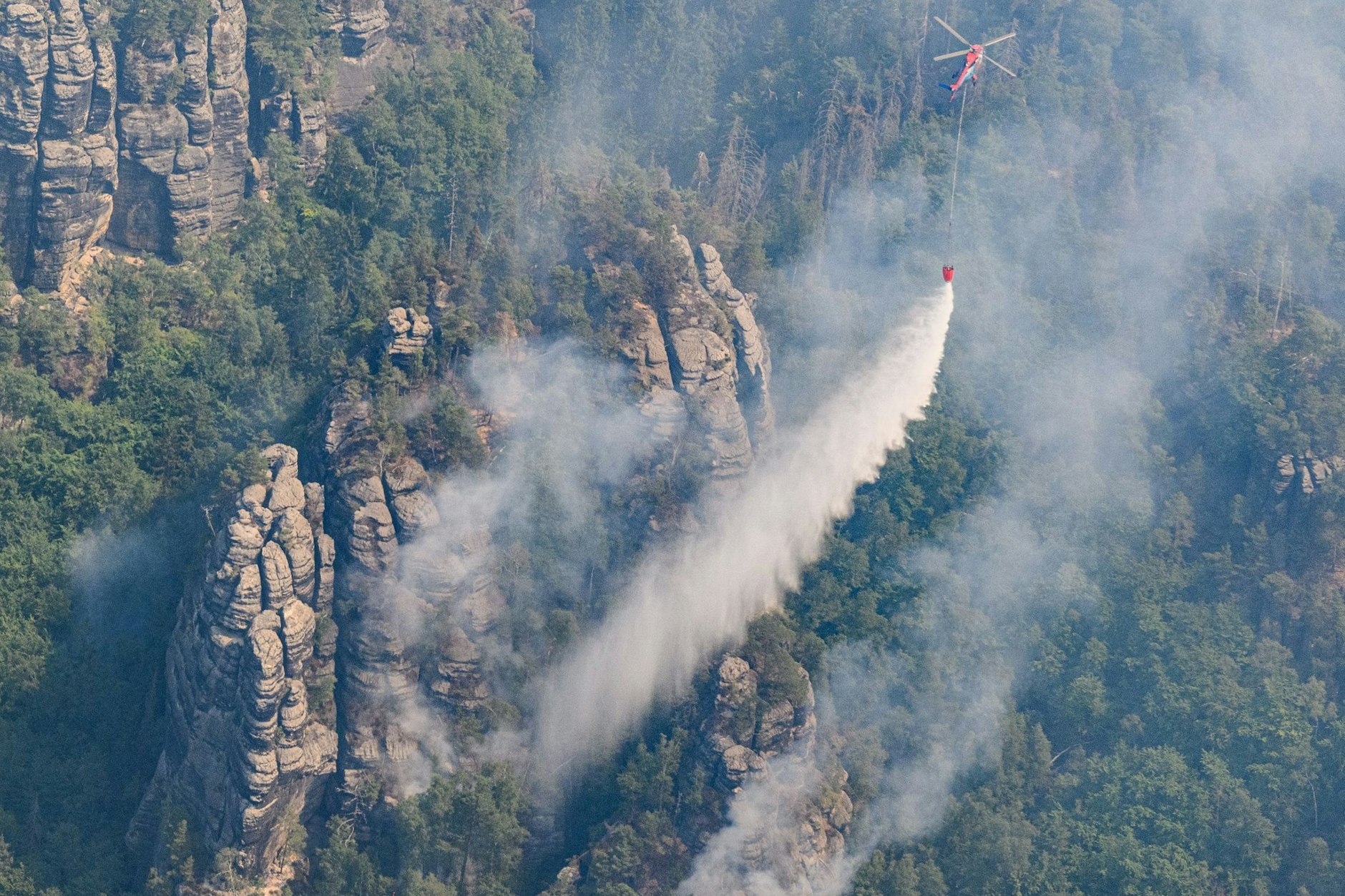Ein Lastenhubschrauber wirft Löschwasser über einem Wald im Nationalpark Sächsische Schweiz ab.