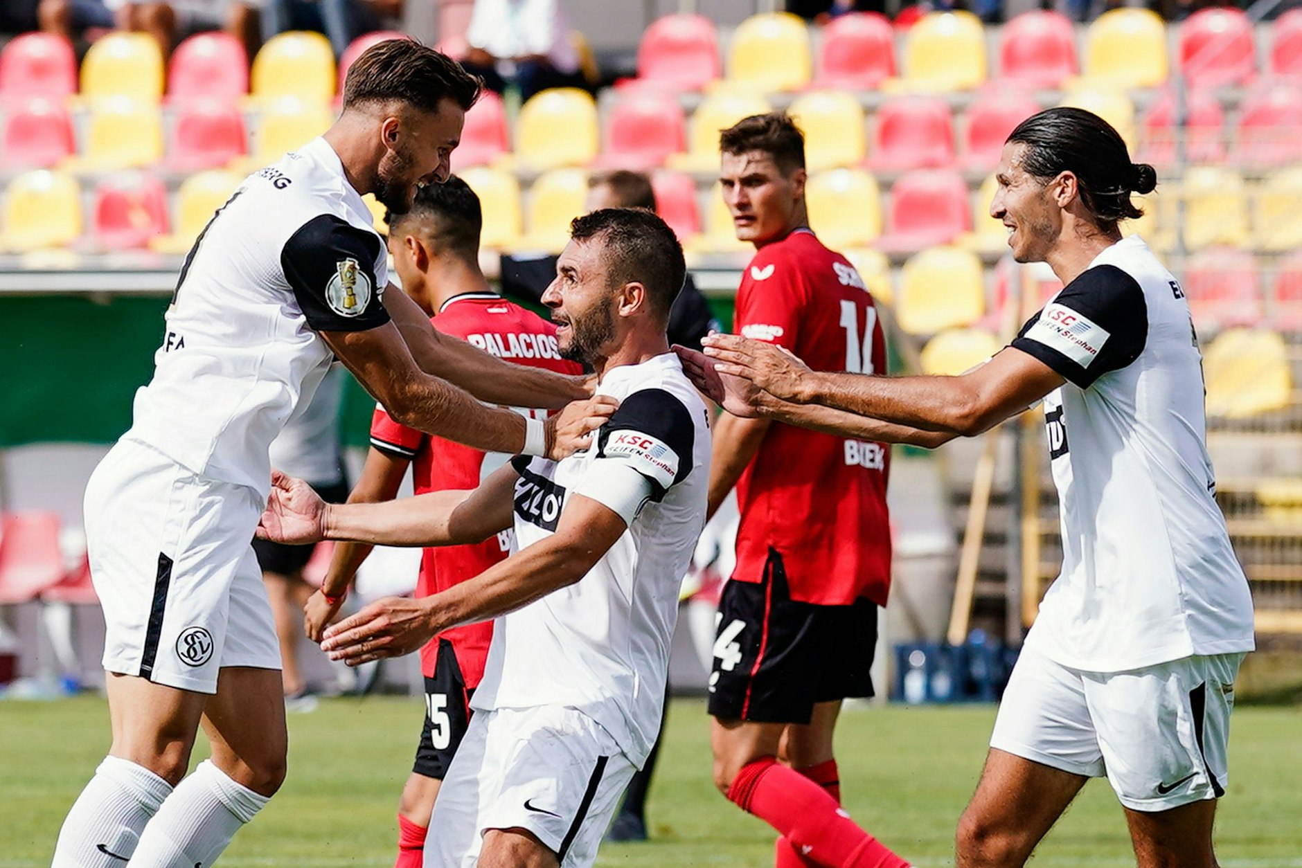 Elversbergs Torschütze Kevin Conrad (M.) von Elversberg jubelt mit den Kollegen. Weiter&nbsp; hinten sieht man eine ziemlich belämmert aussehenden Leverkusener Patrik Schick.