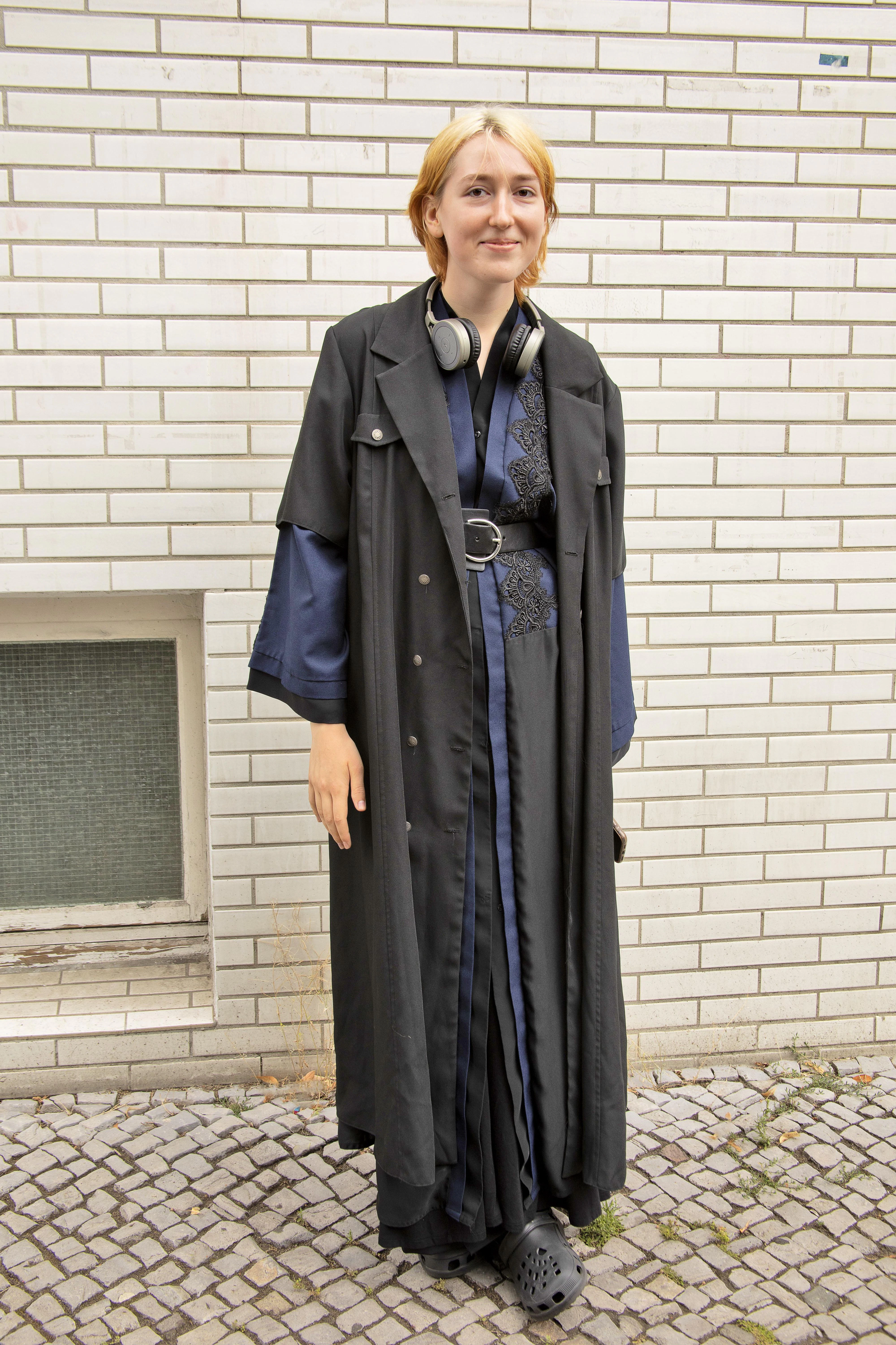 Die Ukrainerin Kateryna war gerade mit der Schule fertig, als der Krieg ausbrach und sie flüchten musste. Hier in Berlin besucht sie jetzt erstmal eine Sprachschule. Ihr raffiniertes Lagenoutfit besteht aus Vintage-Teilen, die sie aus der Heimat mitgebracht hat. Sie erzählt, dass sie keine Sommerschuhe dabei hatte. Also improvisiert sie mit Plastik-Exemplaren in Crocs-Optik.