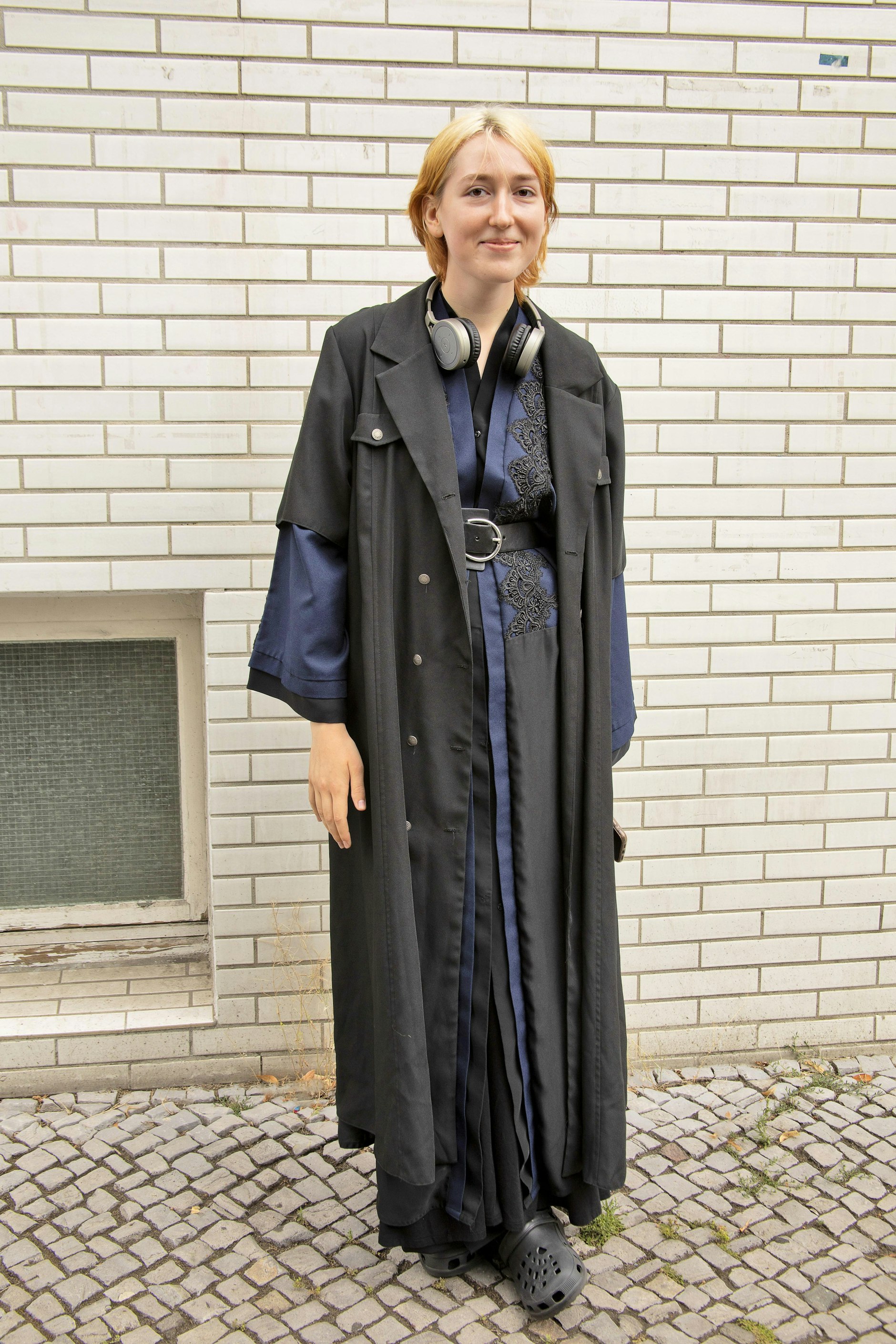 Die Ukrainerin Kateryna war gerade mit der Schule fertig, als der Krieg ausbrach und sie flüchten musste. Hier in Berlin besucht sie jetzt erstmal eine Sprachschule. Ihr raffiniertes Lagenoutfit besteht aus Vintage-Teilen, die sie aus der Heimat mitgebracht hat. Sie erzählt, dass sie keine Sommerschuhe dabei hatte. Also improvisiert sie mit Plastik-Exemplaren in Crocs-Optik.