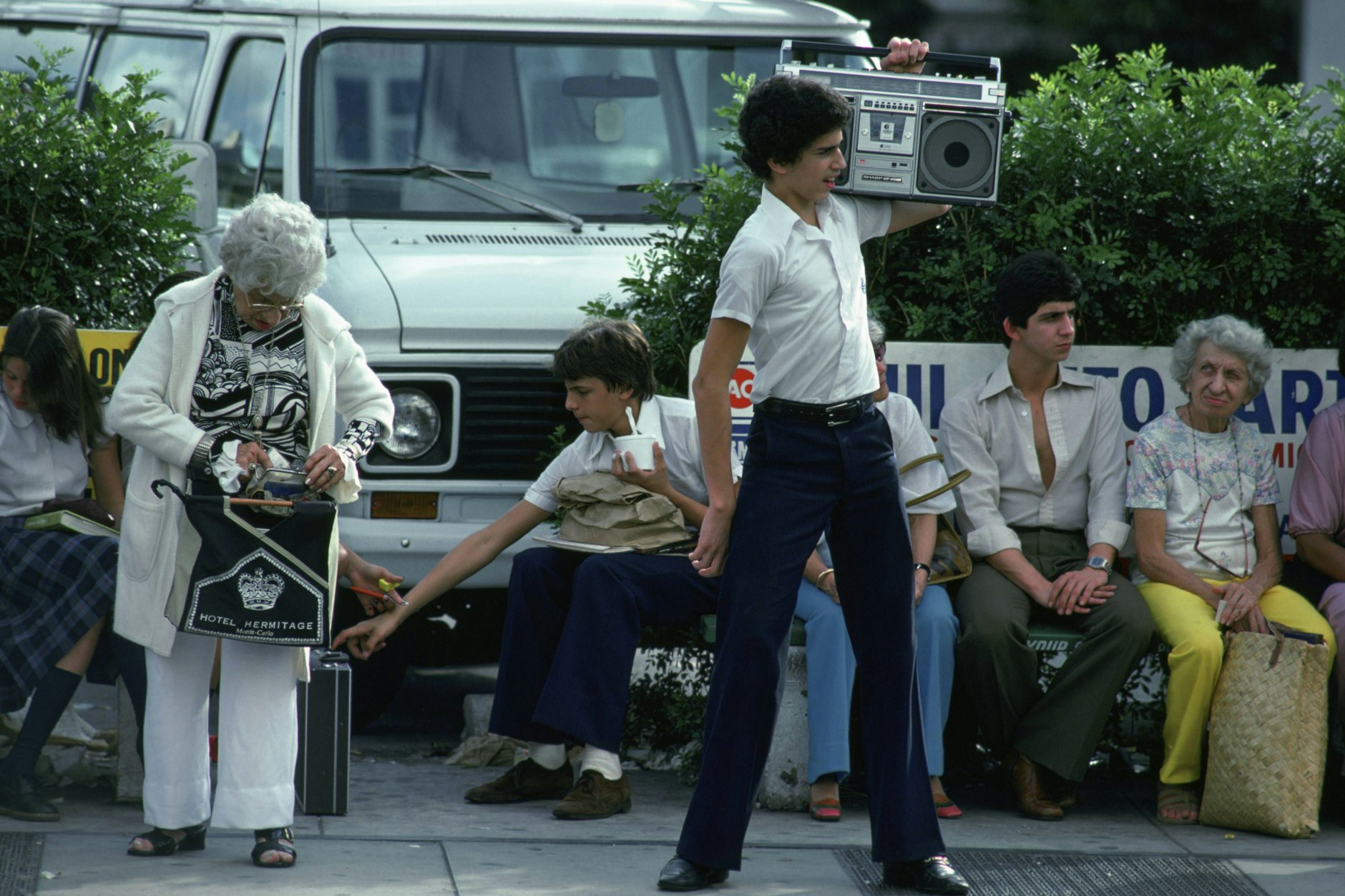 Heute trägt man Airpods, damals eine Boombox: Dieser amerikanische Teenager in Schuluniform beschallt offensichtlich eine Bushaltestelle, wobei das kaum jemanden stört (außer die Dame rechts).
