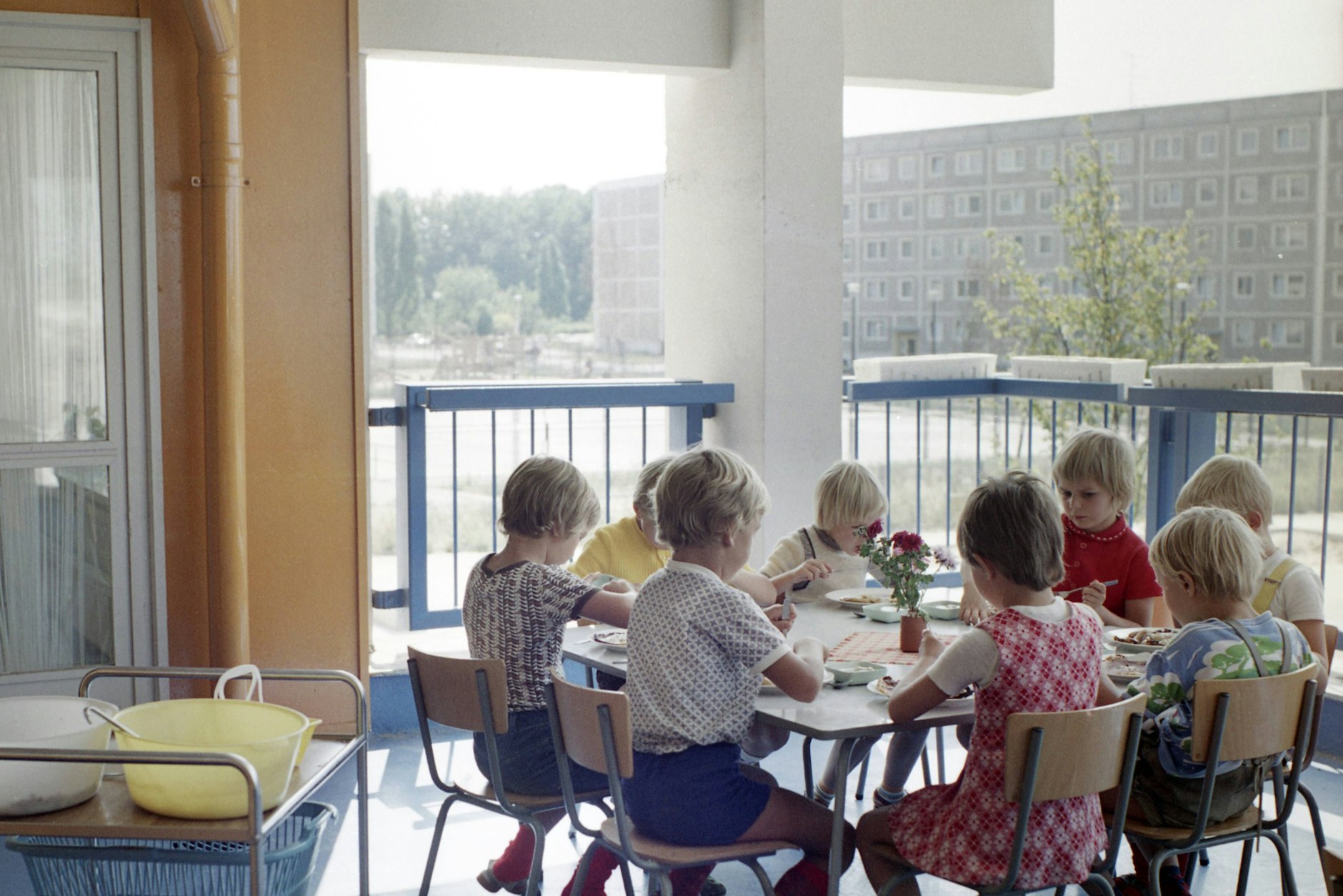 1976: Kinder einer Kindertagesstätte in der Lindenbergallee in Ost-Berlin sitzen zum Essen auf dem Balkon an einem Tisch.