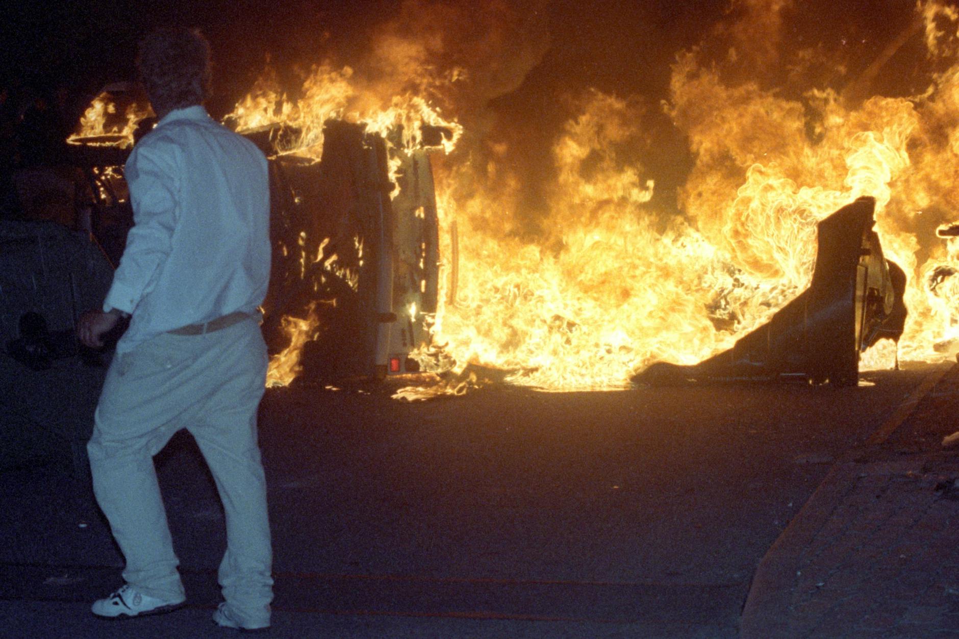 Ein Mann steht am 27.08.1992 vor einem brennenden Pkw auf einer Straße am zentralen Asylbewerberheim in Rostock-Lichtenhagen.