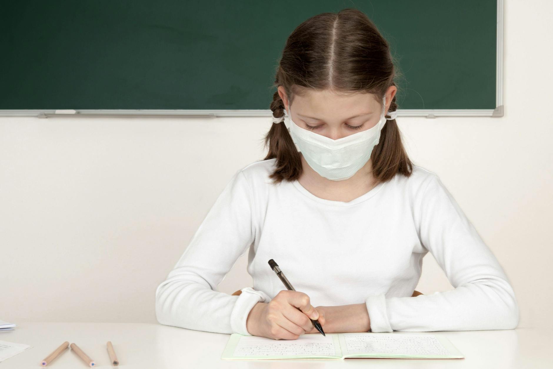 Ein Mädchen mit Maske sitzt an einem Schultisch und schreibt.