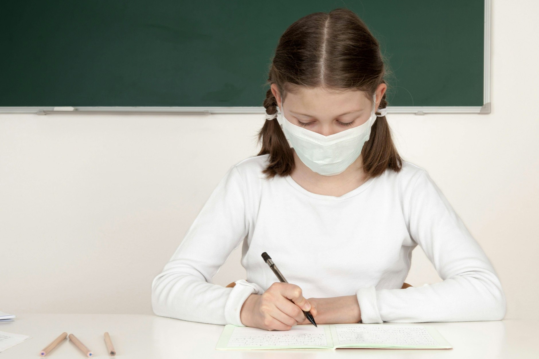 Ein Mädchen mit Maske sitzt an einem Schultisch und schreibt.