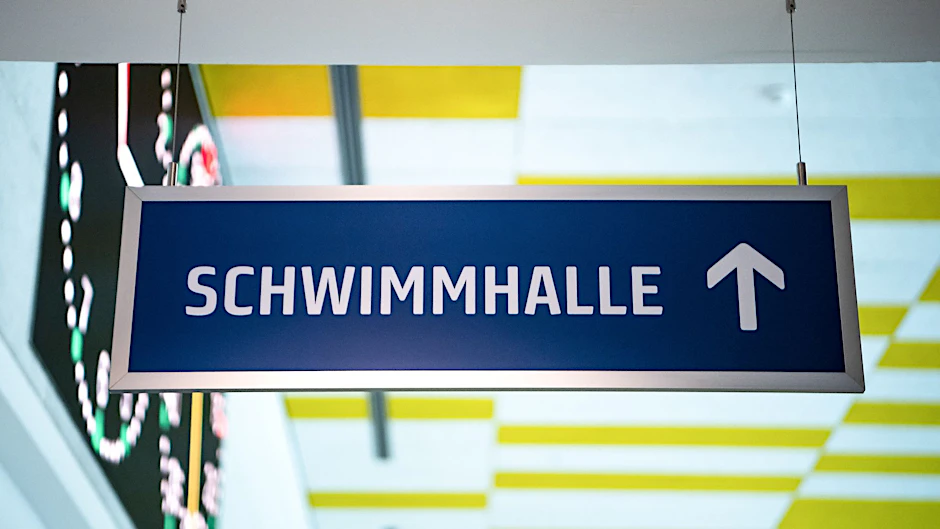 PRODUKTION - Ein Schild weißt zu einer Schwimmhalle.  