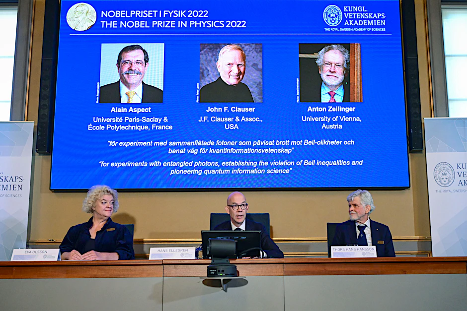 Bekanntgabe des Physik-Nobelpreises 2022 in Stockholm. Auf dem Bildschirm oben von links nach rechts: Alain Aspect, John F. Clauser und Anton Zeilinger.