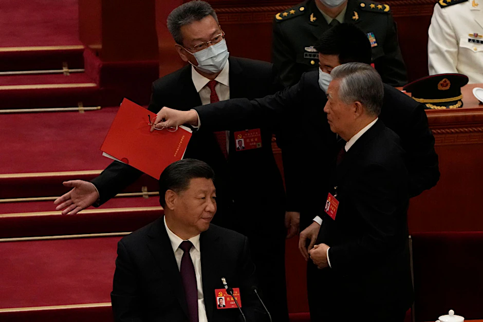 Chinas Ex-Präsident Hu Jintao (r) wird von seinem Platz weg begleitet. Links sitzt Xi Jinping und guckt zu.  