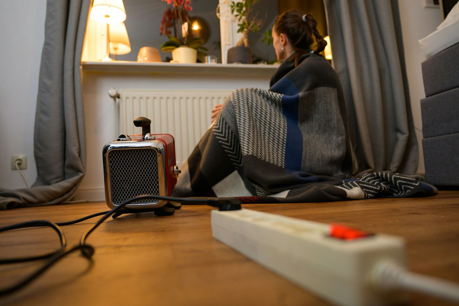 Eine Frau sitzt mit einer Decke hinter einem Elektro-Heizlüfter.