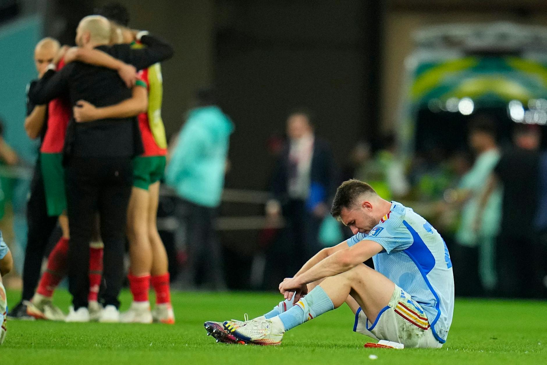 Spaniens Aymeric Laporte sitzt nach der Niederlage auf dem Spielfeld, während Marokkos Spieler im Hintergrund über ihren Sieg im Elfmeterschiessen jubeln.
