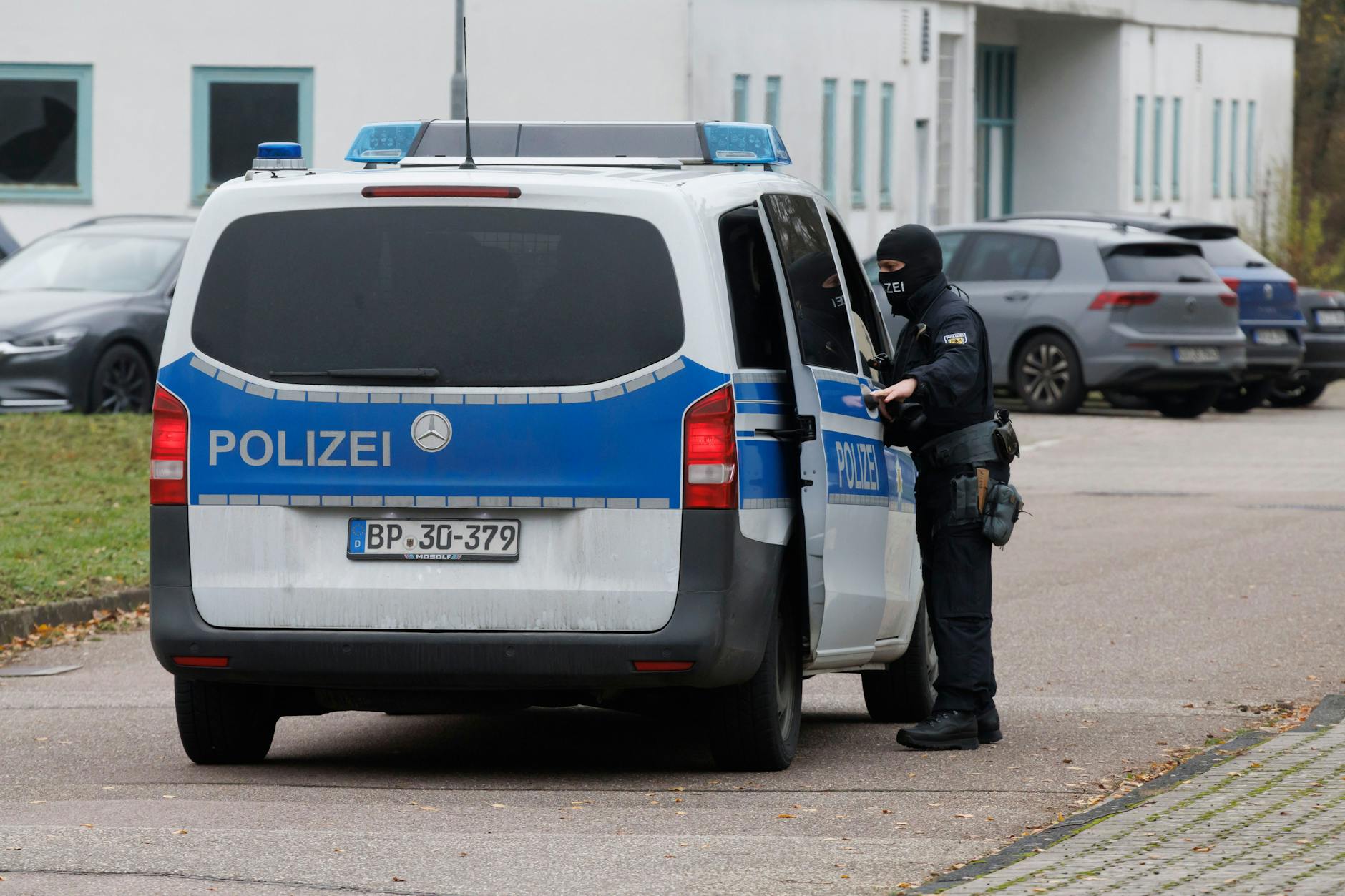 An einer Außenstelle vom Bundesgerichtshof fährt ein Streifenwagen mit Beschuldigten vor. Ein vermummter Polizist öffnet die Schiebetür. 