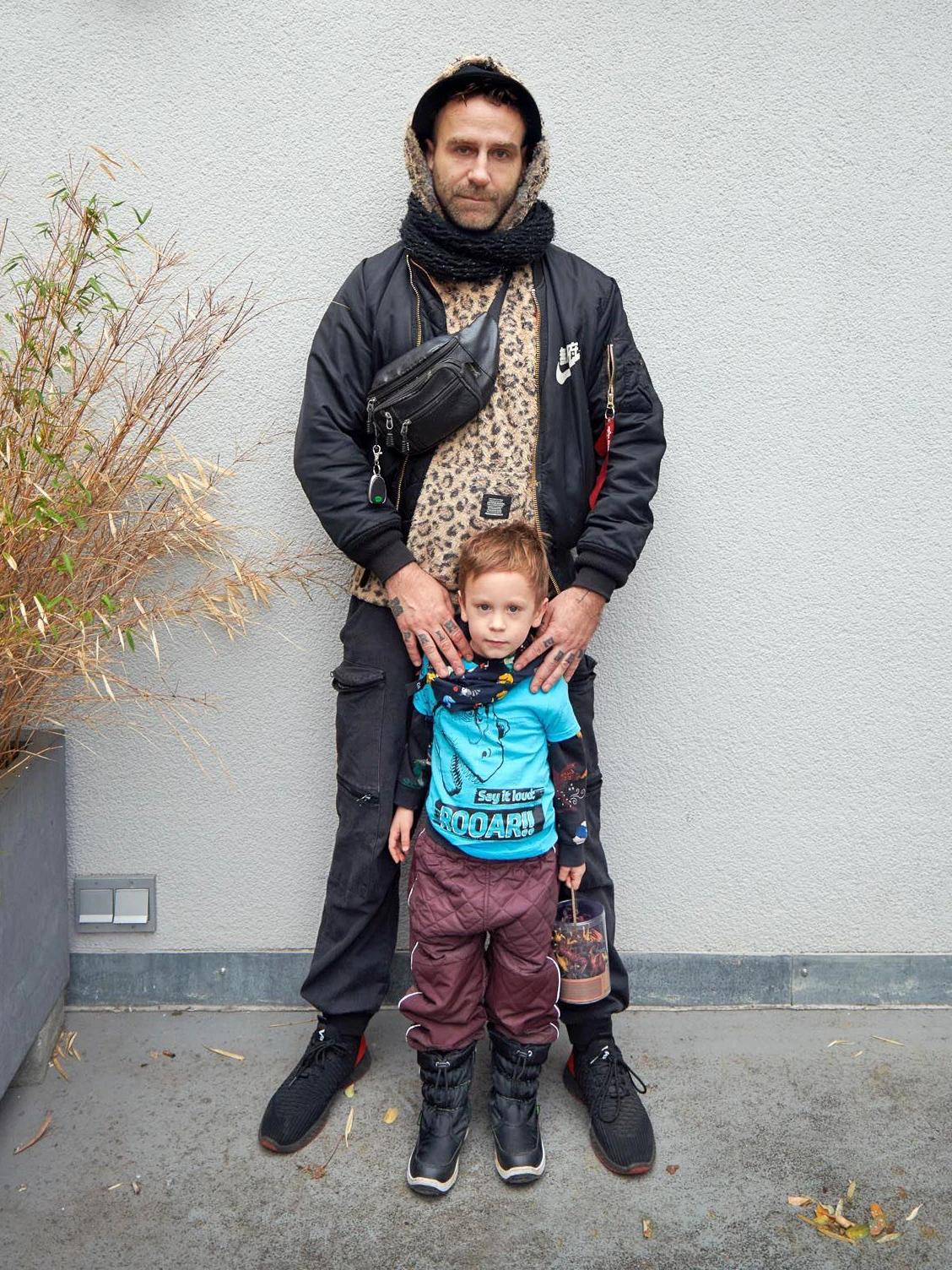 Der dreifache Vater Jan ist mit seinem Sohn Louis unterwegs. Jan ist DJ und Veranstalter der Fetisch-Partyreihe „Kinky Galore“, die er vor zehn Jahren in Berlin ins Leben rief. Er hat sein Schlechtwetteroutfit an, wie er sagt: Unter der Bomberjacke (Alpha Industries x Nike) einen Flauschpullover mit Leo-Print, dazu eine schwarze Cargo-Hose und No-name-Sneaker. Der vierjährige Louis macht gerade eine Dino-Phase durch, entsprechend sind Saurier auf T-Shirt und Halstuch zu sehen.