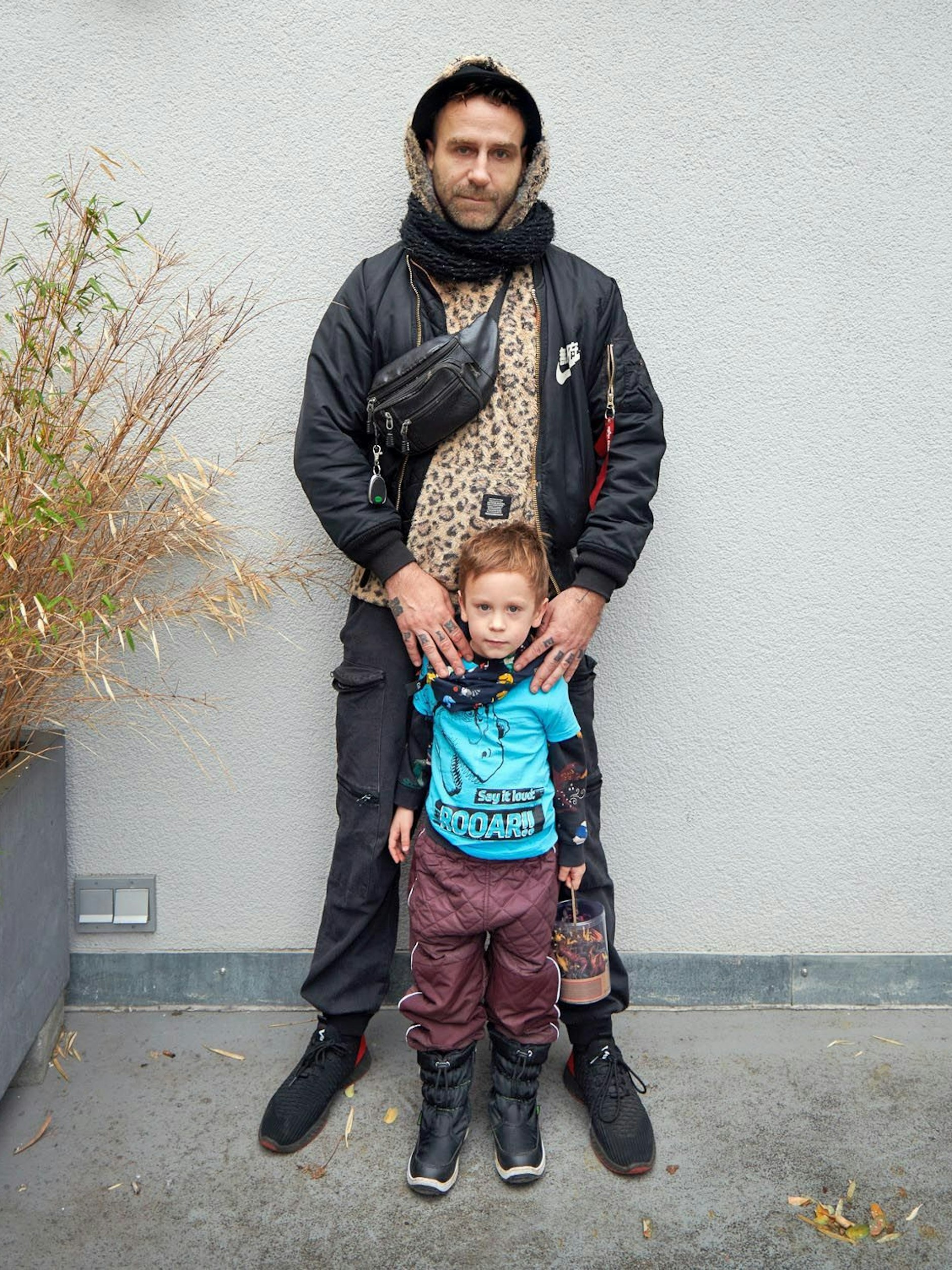 Der dreifache Vater Jan ist mit seinem Sohn Louis unterwegs. Jan ist DJ und Veranstalter der Fetisch-Partyreihe „Kinky Galore“, die er vor zehn Jahren in Berlin ins Leben rief. Er hat sein Schlechtwetteroutfit an, wie er sagt: Unter der Bomberjacke (Alpha Industries x Nike) einen Flauschpullover mit Leo-Print, dazu eine schwarze Cargo-Hose und No-name-Sneaker. Der vierjährige Louis macht gerade eine Dino-Phase durch, entsprechend sind Saurier auf T-Shirt und Halstuch zu sehen.