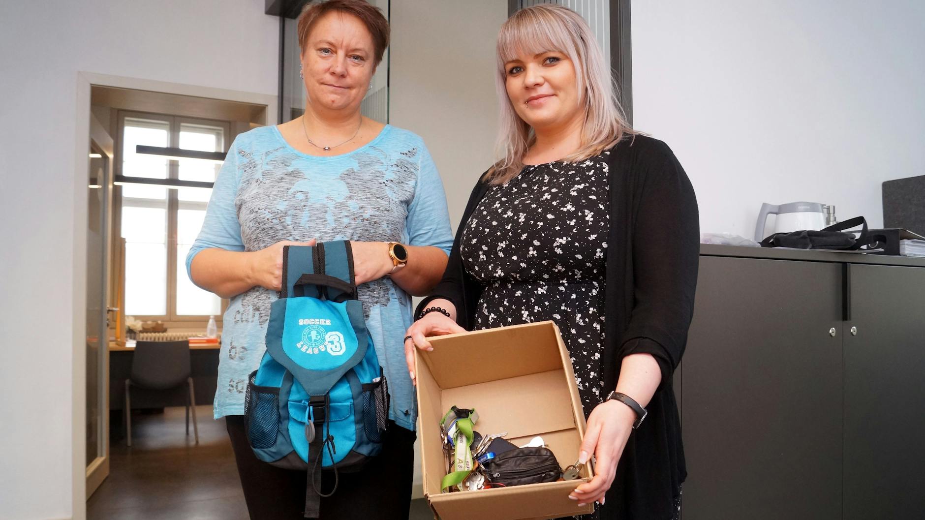 Peggy Samusch (l.) und Claudia Schulze vom Wittstocker Fundbüro mit einem Kinder-Rucksack und einem Karton mit verbummelten Schlüsseln.