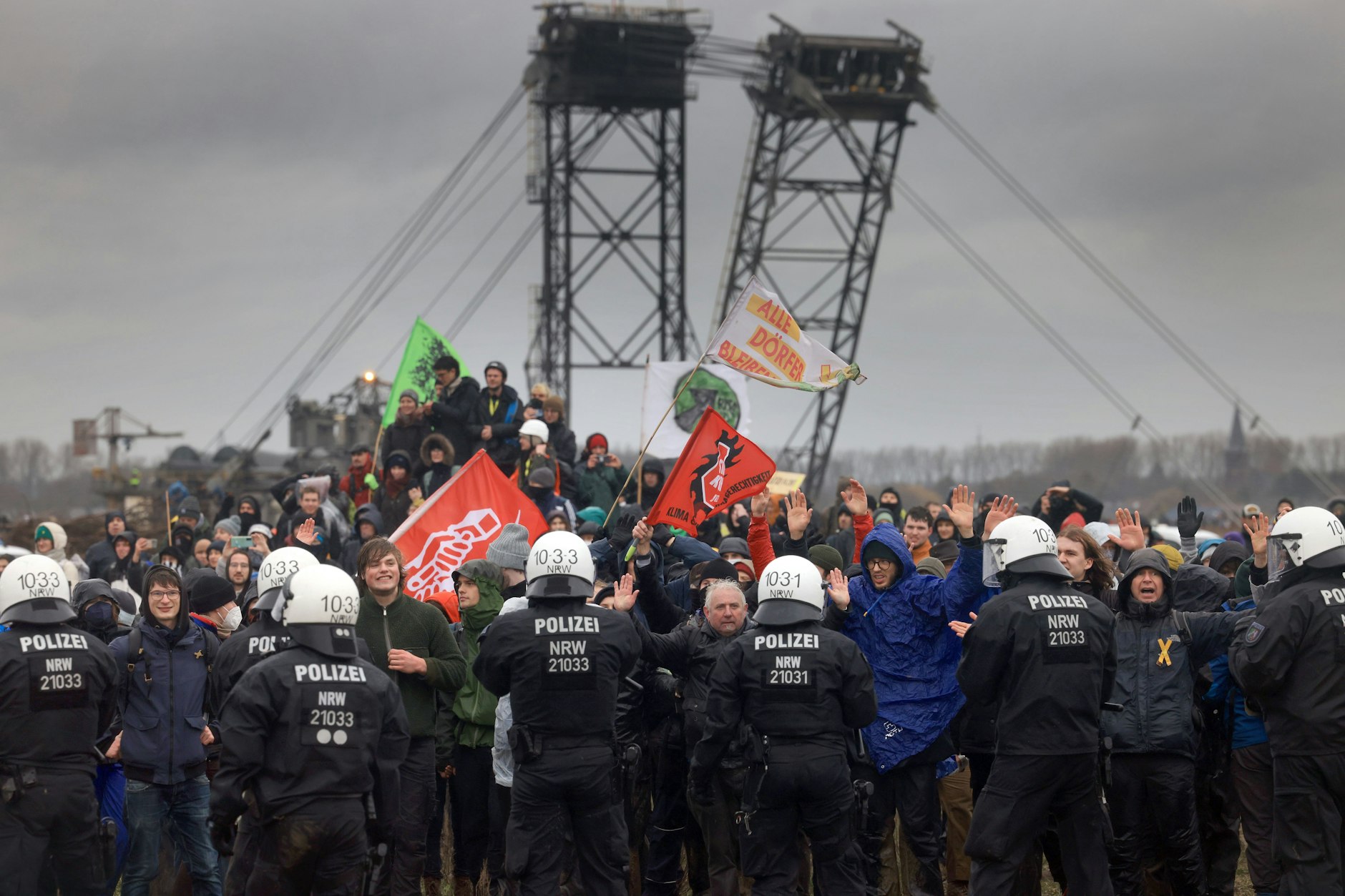 Polizisten und Demonstranten mit erhobenen Händen stehen sich bei der Demonstration von Klimaaktivisten am Rande des Braunkohletagebaus bei Lützerath gegenüber, im Hintergrund ein Kohlebagger.