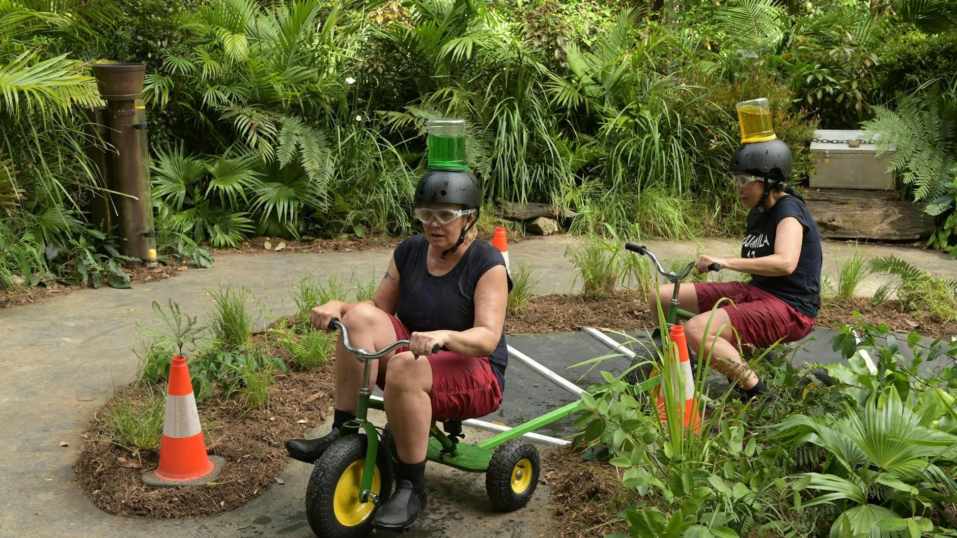 Claudia Effenberg (l.) und Djamila Rowe müssen bei ihrer Dschungelcamp-Schatzsuche Flüssigkeiten in Behältern auf ihrem Kopf zu einem Topf befördern.