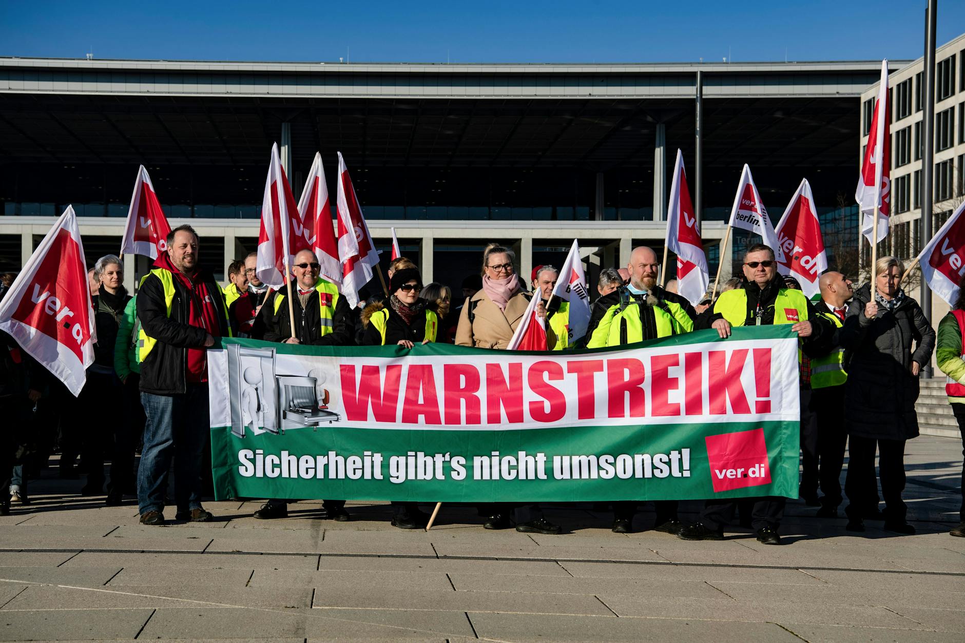 „Warnstreik“ steht auf dem Transparent, hinter dem sich Mitarbeiter des Sicherheitspersonals am BER versammelt haben, am 22.3.2022.